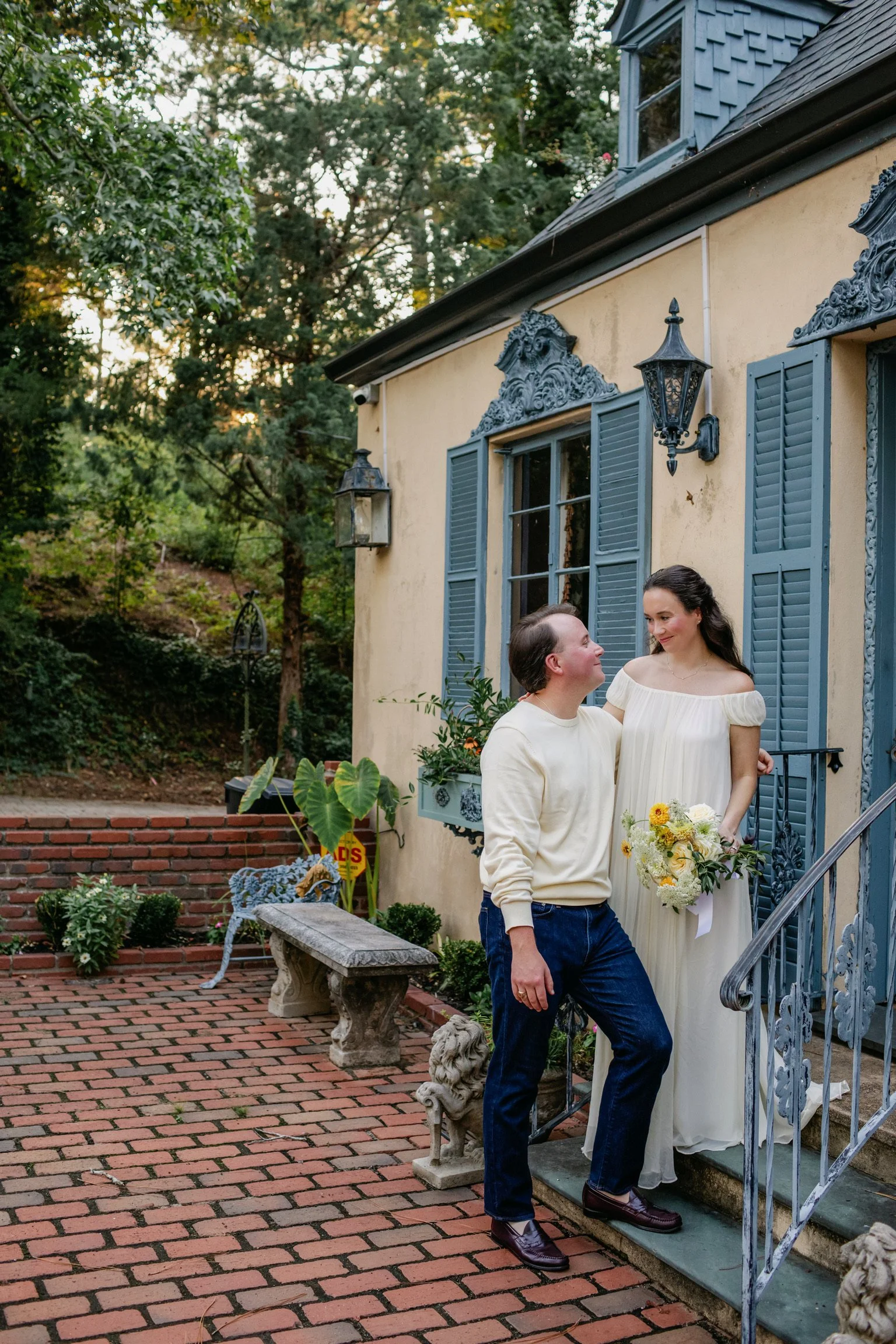 A couple standing on brick stairs outside a house with light yellow walls and blue shutters, the woman wearing a white dress and holding a bouquet of flowers, the man in a cream sweater and dark jeans, both smiling at each other, surrounded by greenery and outdoor decor.