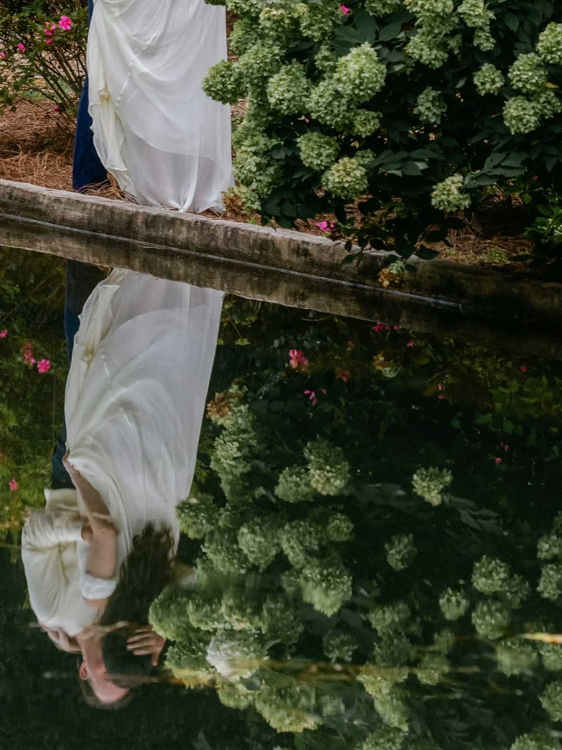 A woman dressed in a white dress standing near a pond with her reflection visible in the water, surrounded by green foliage and pink flowers.