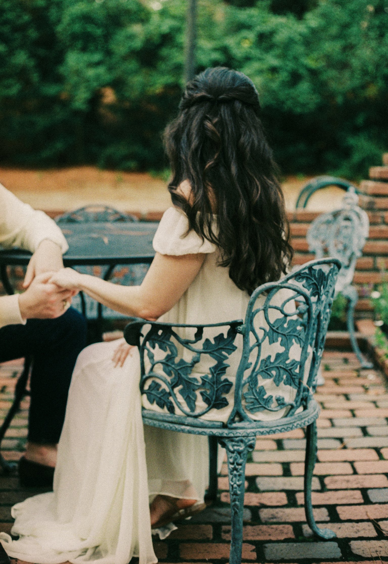 A woman in a cream-colored dress sitting on a blue ornate metal chair at an outdoor patio, holding hands with someone across from her, with trees and a brick wall in the background.