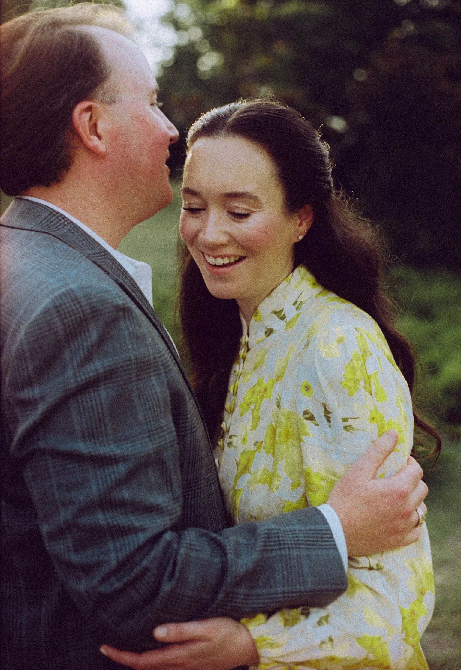 A man and woman dressed formally, embracing outdoors with trees in the background, the woman smiling with eyes closed.