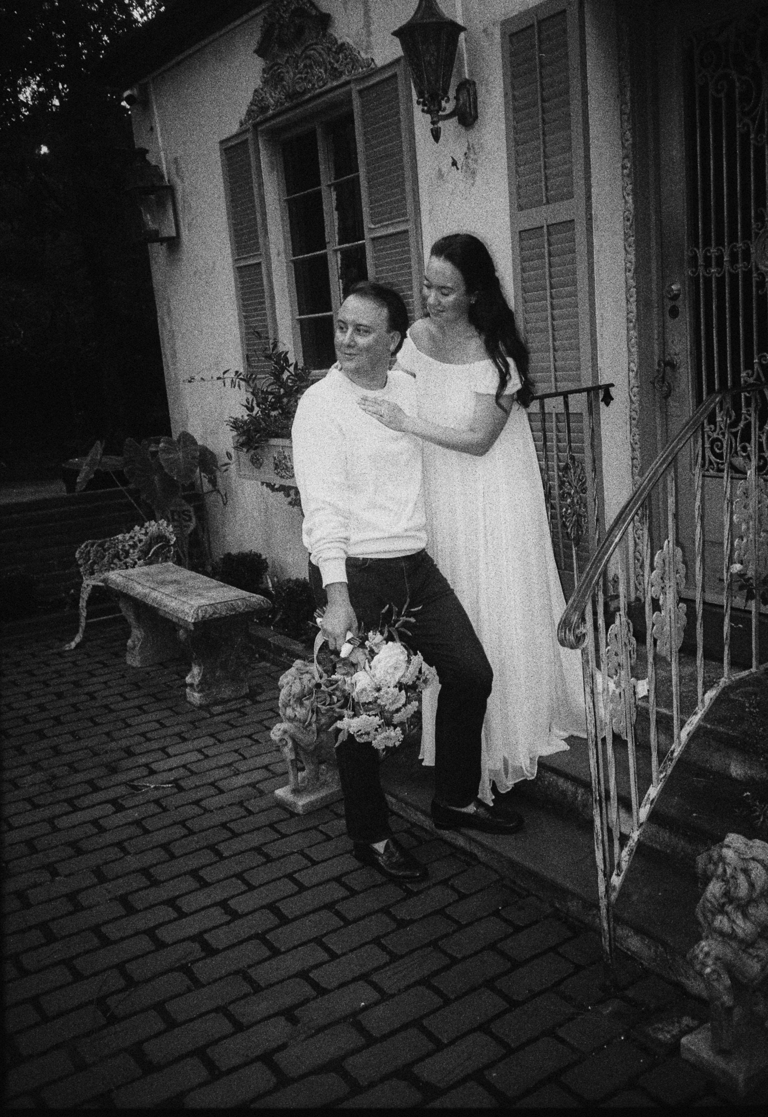 A man and a woman standing together on the steps of a house, with the woman in a white dress and the man holding a bouquet of flowers.