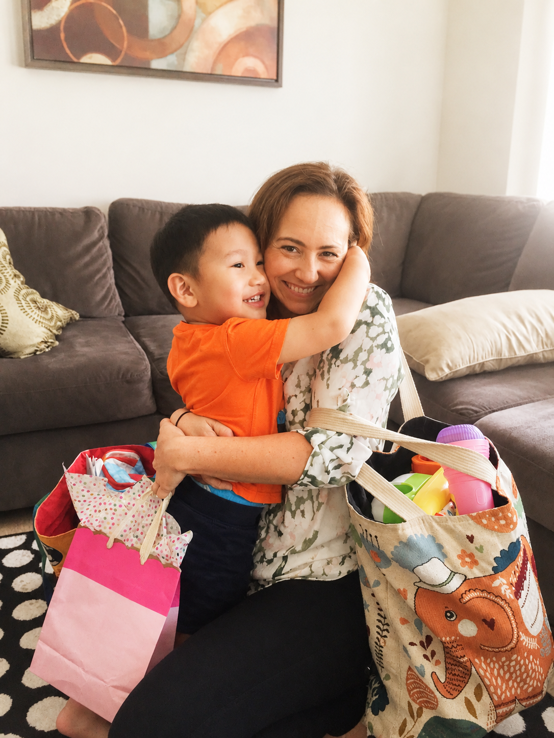 A woman and young boy hugging each other inside a living room. The woman is holding bags of toys and a pink gift bag. The boy is wearing an orange shirt and black shorts, smiling and hugging the SLP on his graduation day in California.