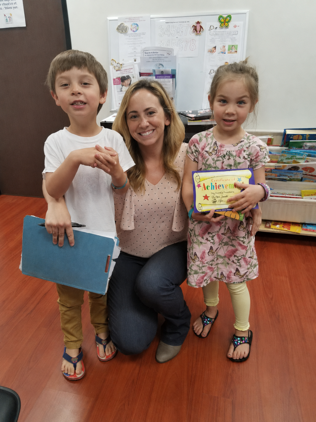 A woman SLP with blonde hair and two children, a boy and a girl, standing in a classroom with books and a whiteboard in the background. The girl is holding a colorful certificate of achievement.