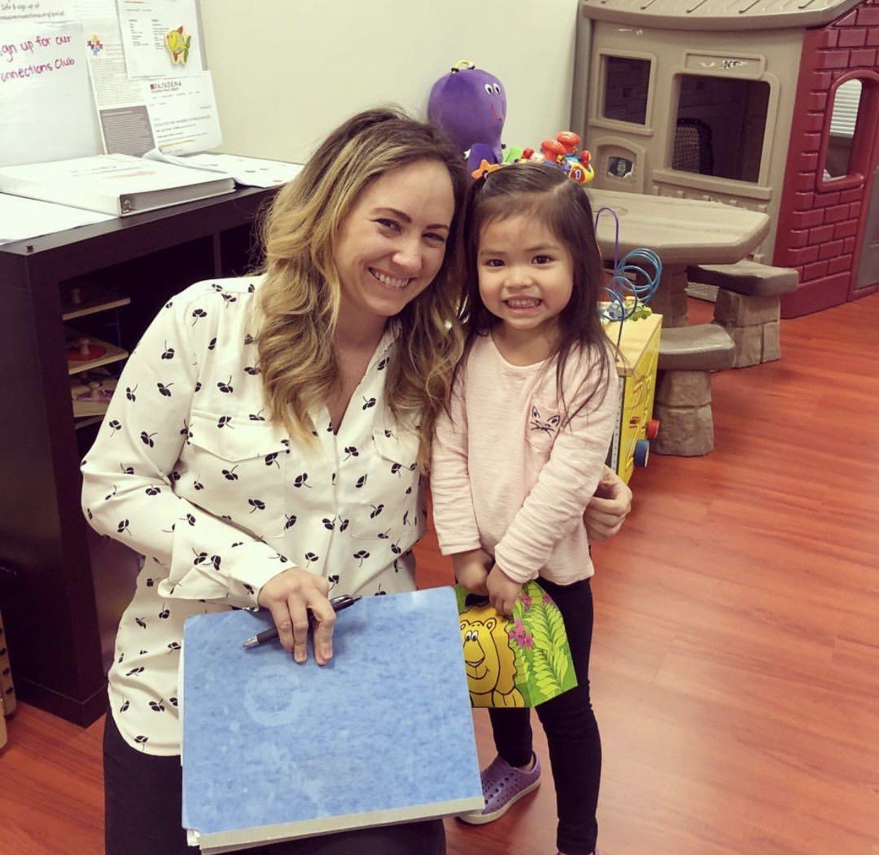 A woman and a young girl smiling and posing together in a playroom. The woman is holding a blue notebook and a pen, and the girl is holding a colorful book with a lion on the cover. There are children's toys and a play kitchen in the background.