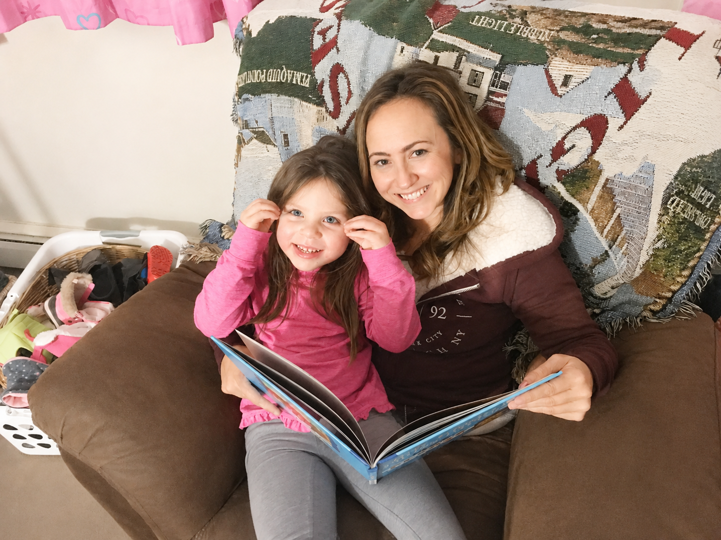A woman and young girl sitting on a brown couch, smiling at the camera while reading a book. The girl is wearing a pink long-sleeve shirt, and the woman is wearing a brown jacket with a white, fuzzy lining. 