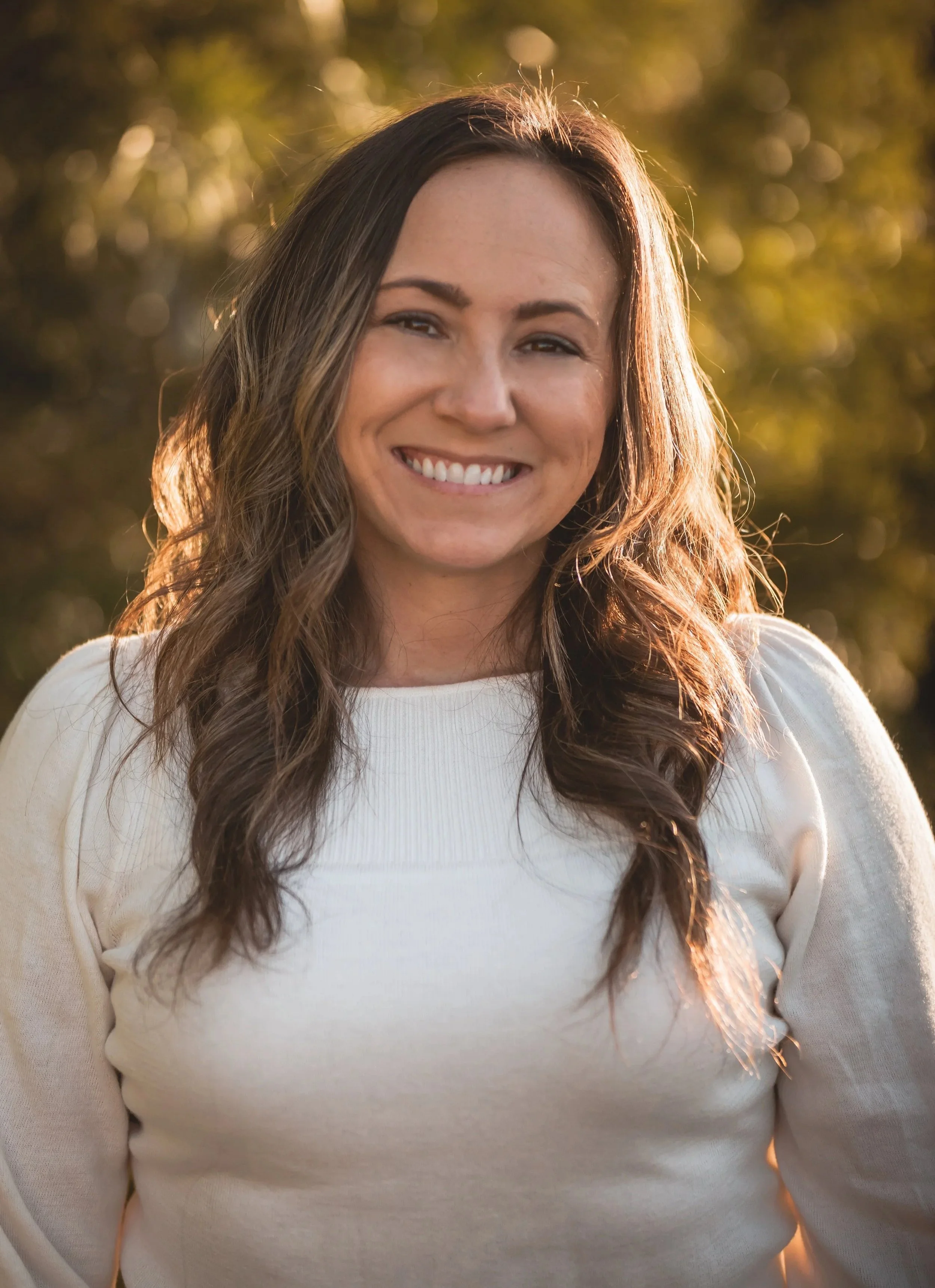 Close-up of a smiling woman with long, wavy brown hair, wearing a cream-colored sweater, outdoors during golden hour.