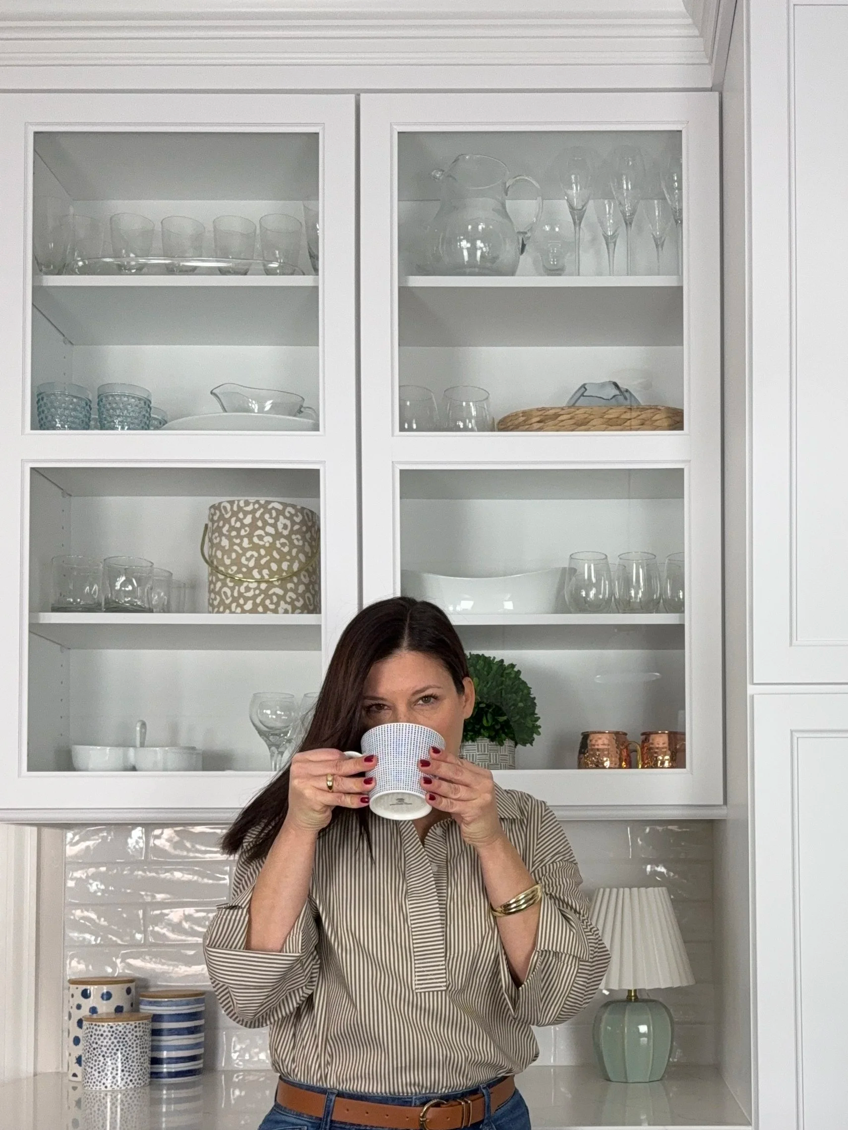 A woman with dark hair and a striped beige and black shirt drinks from a white mug in a white kitchen with glassware on shelves and decorative kitchenware on the counter.