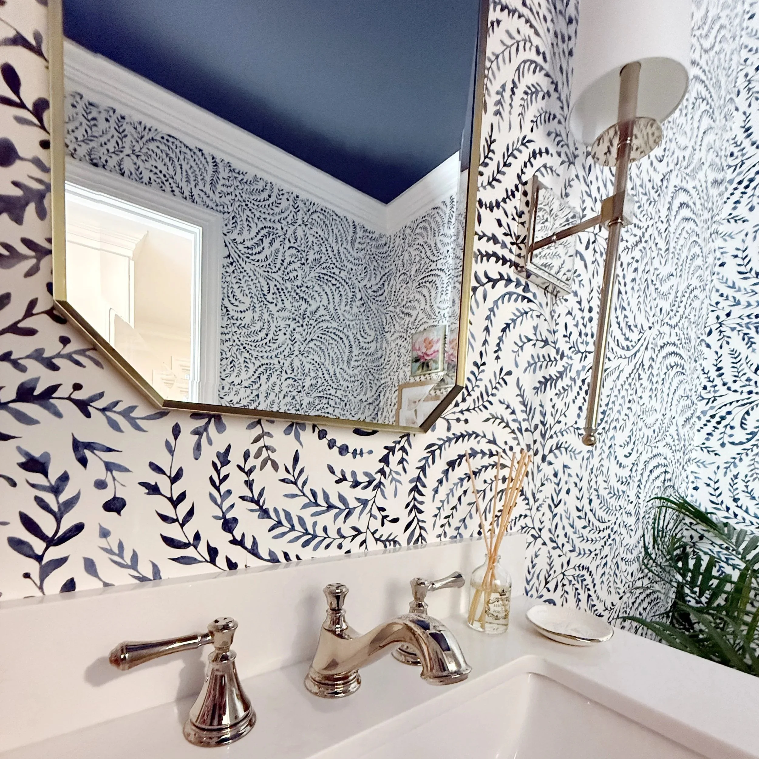 Close-up of a bathroom sink with a silver faucet, mirror, decorative wallpaper with blue leaves, and a small framed picture of flowers in the background.