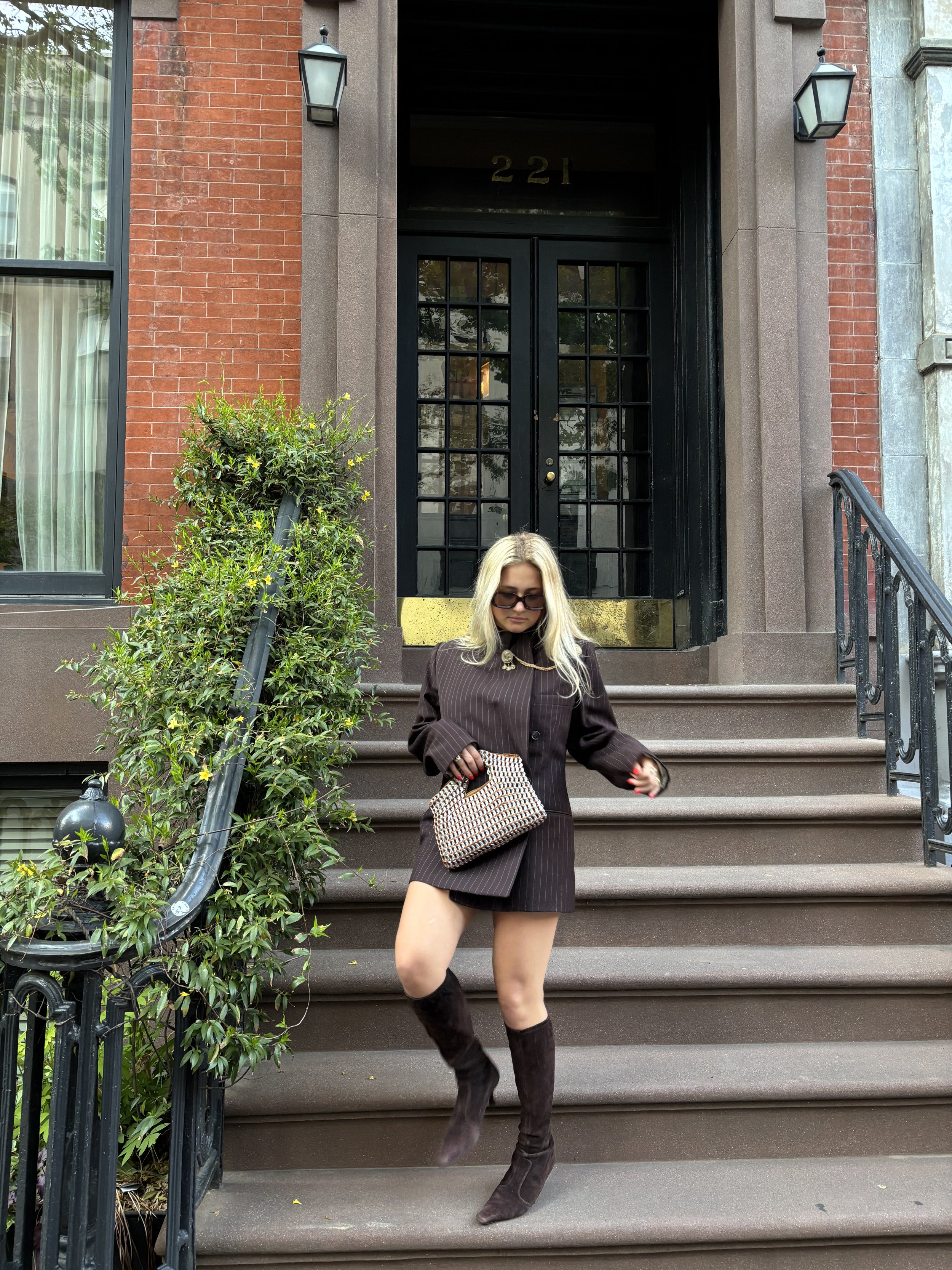 A woman with long blonde hair, wearing sunglasses, a dark pinstripe blazer, a matching skirt, and over-the-knee brown boots, standing on the steps of a brick building with black doors and large windows, carrying a checkered handbag.