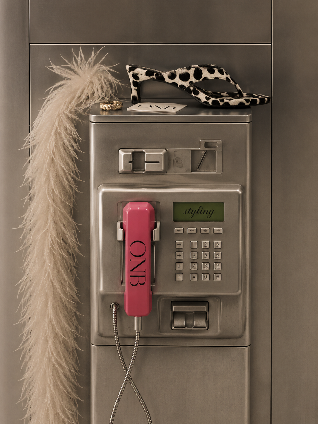 A vintage metal payphone with a pink handset labeled 'ONB', decorated with a feathered accessory and a pair of black-and-white polka-dot high-heeled shoes on top. A gold ring and a small card reading 'ONE' rest on the nearby surface.
