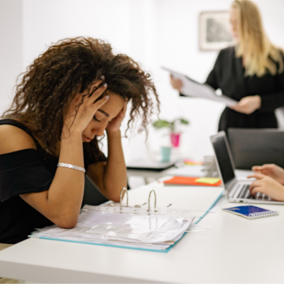 A woman with curly hair sitting at a desk, holding her head in frustration surrounded by paperwork, with a woman in the background holding a folder.