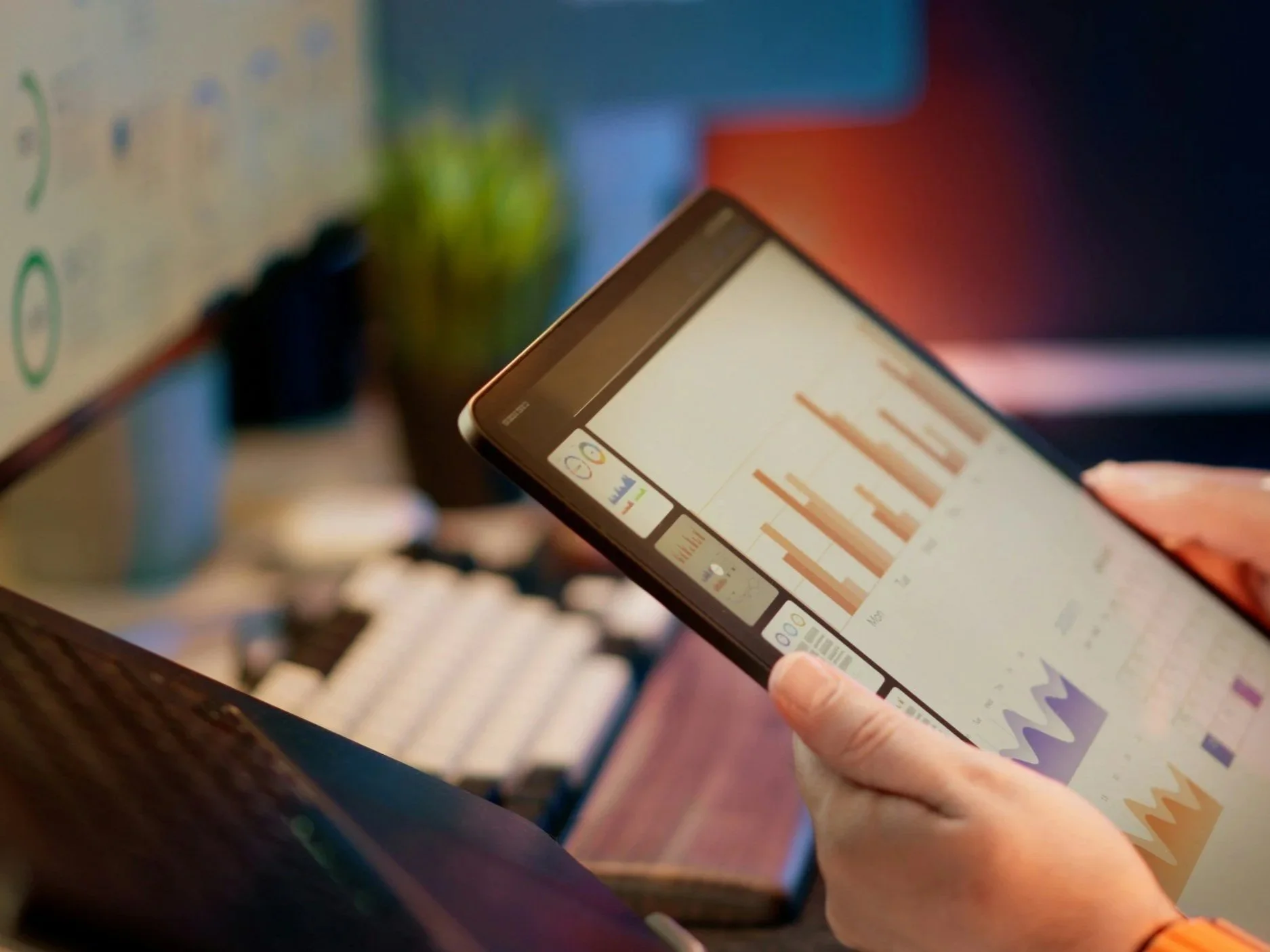 Person holding a tablet displaying graphs and data charts, with a blurry background showing a computer monitor and office environment.