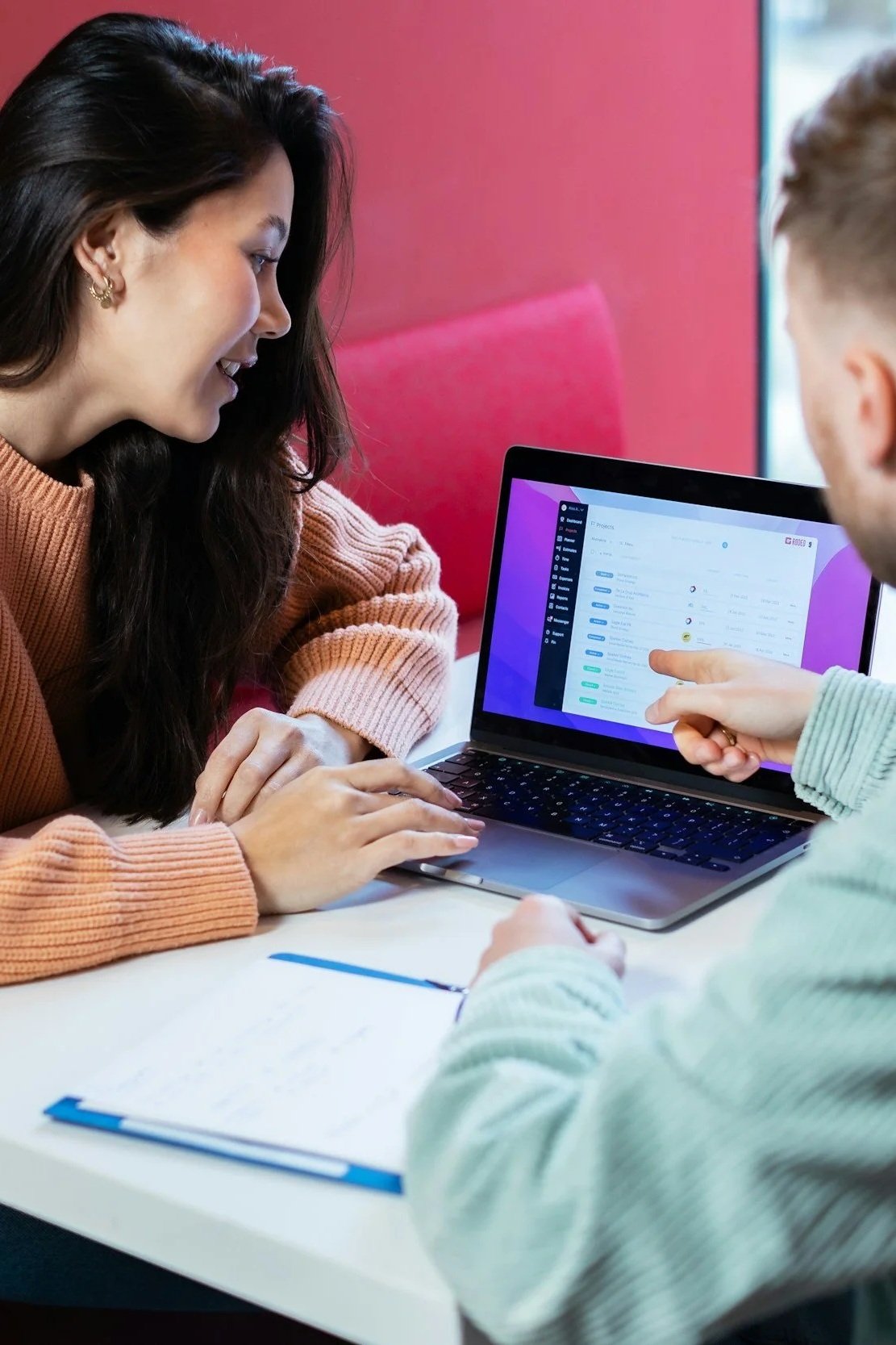 Two people reviewing a project management software on a laptop at a meeting table, with notes and pens nearby.