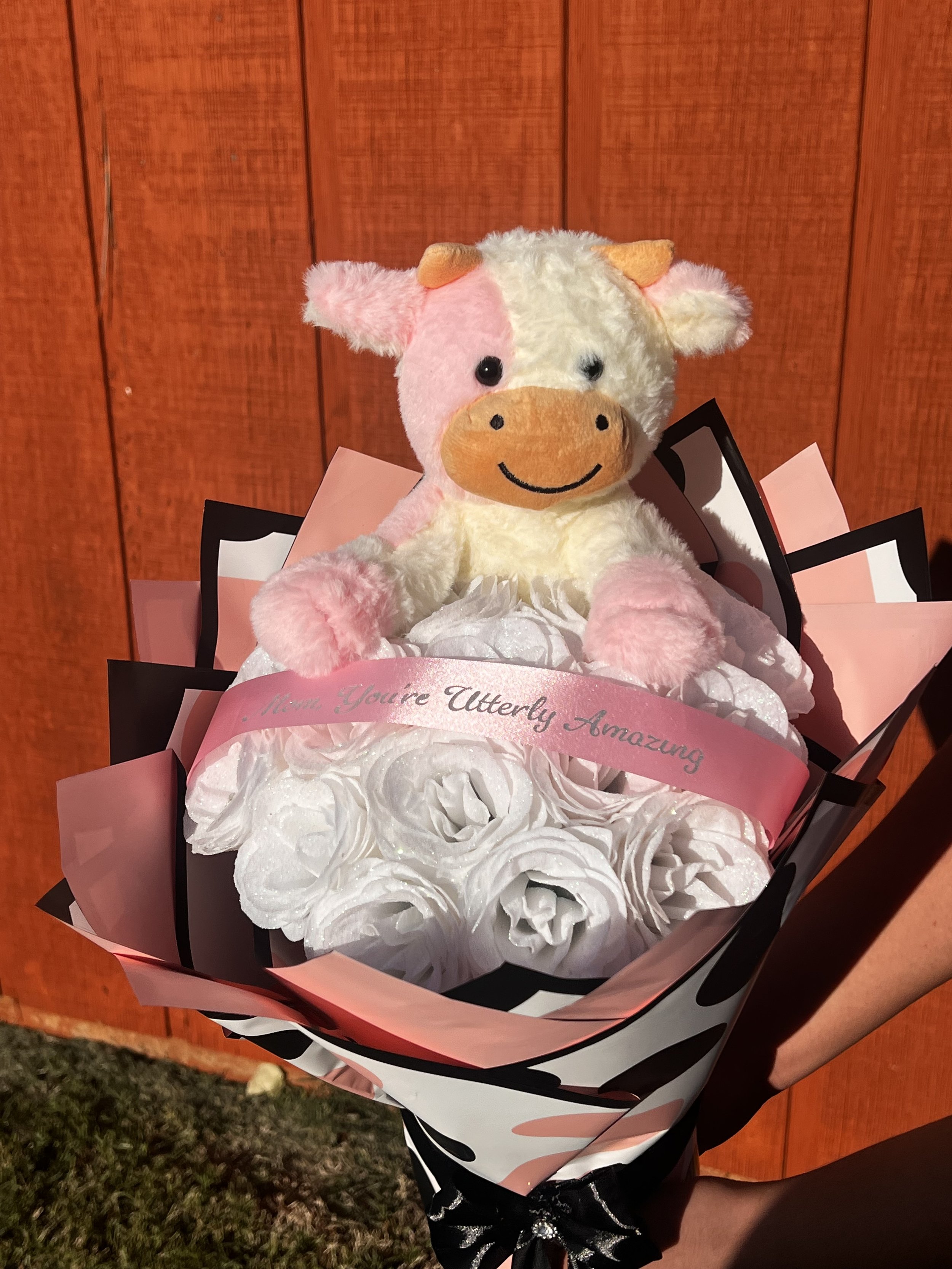A stuffed plush pig toy is sitting on top of a bouquet of white flowers, held by a person with a black dress with silver sparkles. The bouquet has a pink ribbon with the message 'You're Utterly Amazing.' The background is a wooden wall.