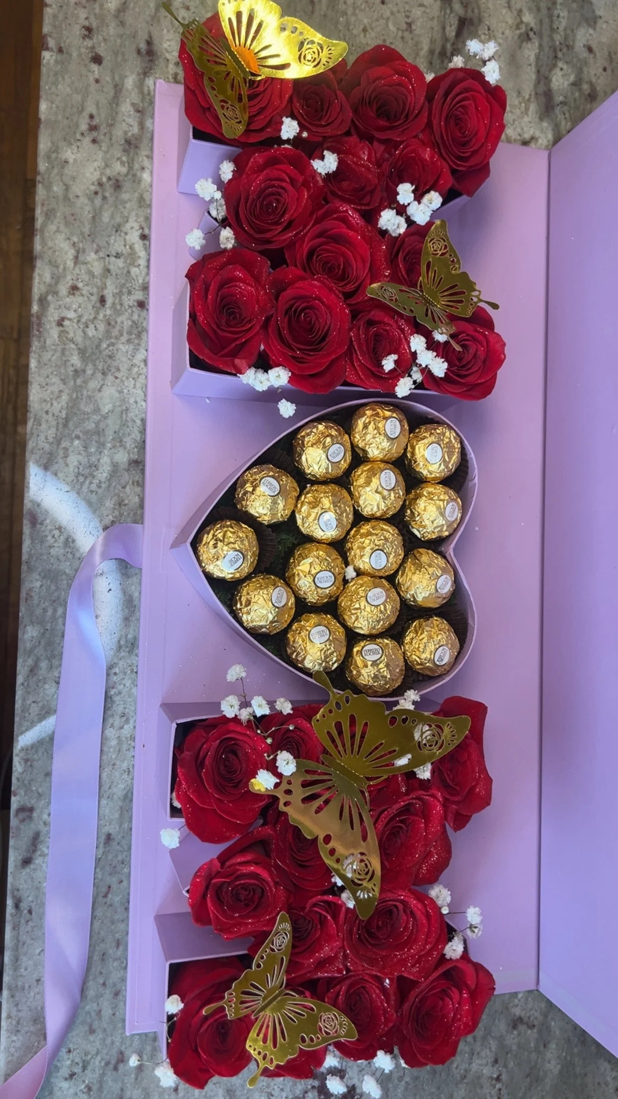 A pink box with red roses, gold butterfly decorations, white baby's breath flowers, and a heart-shaped box of Ferrero Rocher chocolates on a purple surface.
