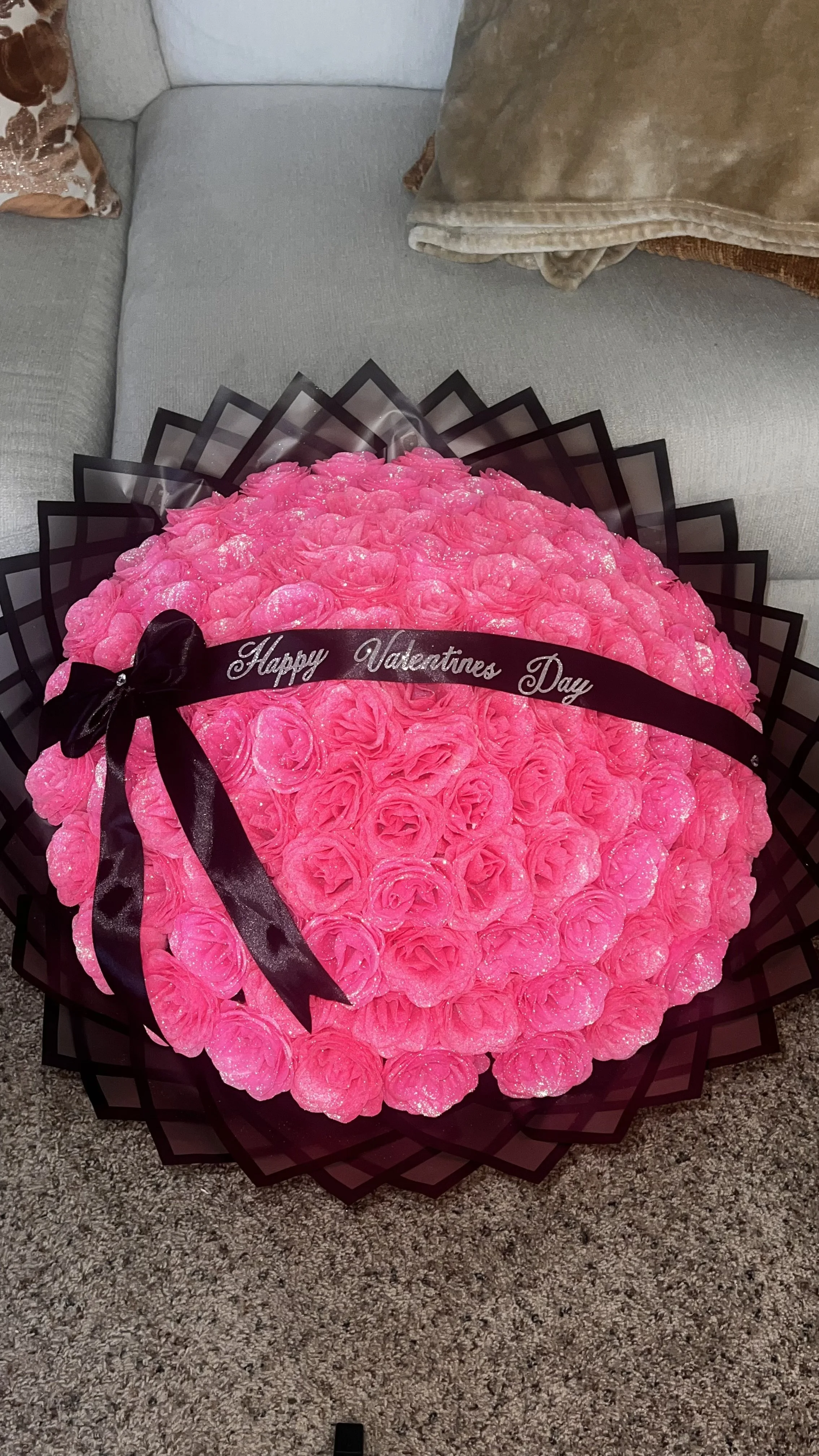 A large bouquet of pink roses with a black ribbon that says 'Happy Valentines Day' resting on a beige sofa with plush cushions.