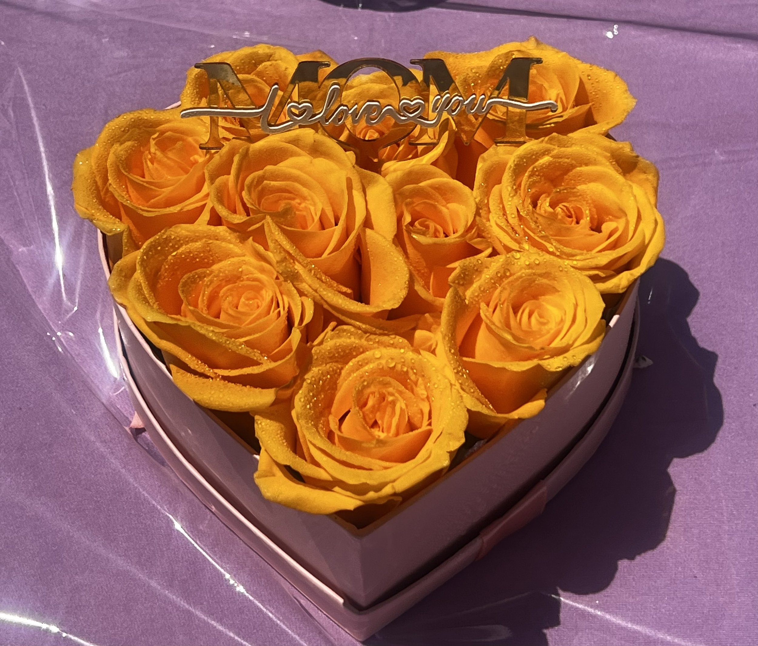 A bouquet of yellow roses with water droplets, adorned with a metallic 'Love You Mom' sign, resting on a purple surface.