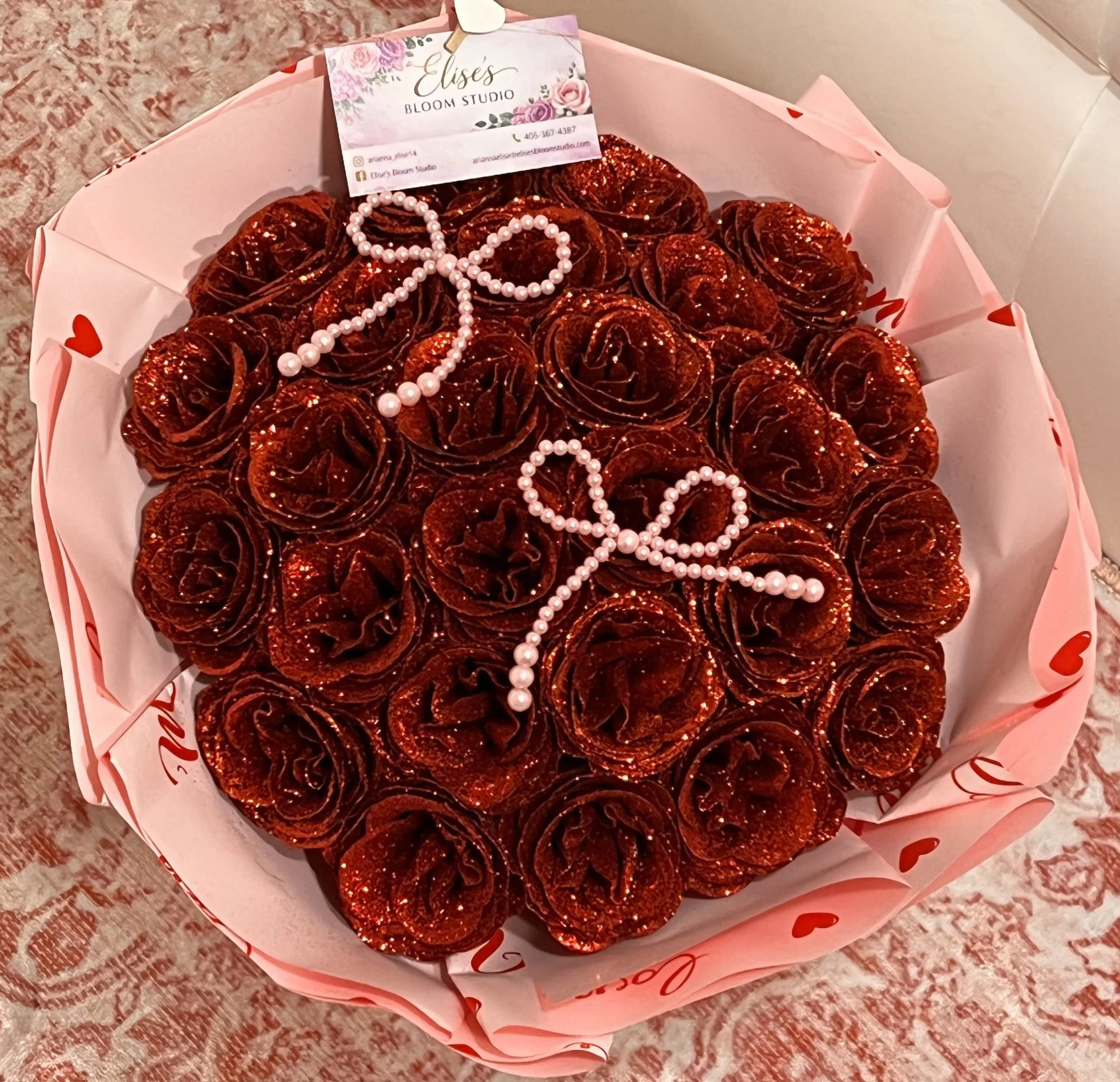 A bouquet of red, glittered caramel apple-shaped treats decorated with pearl necklaces and a business card from Elise's Bloom Studio, placed inside a pink paper wrapping with heart designs.