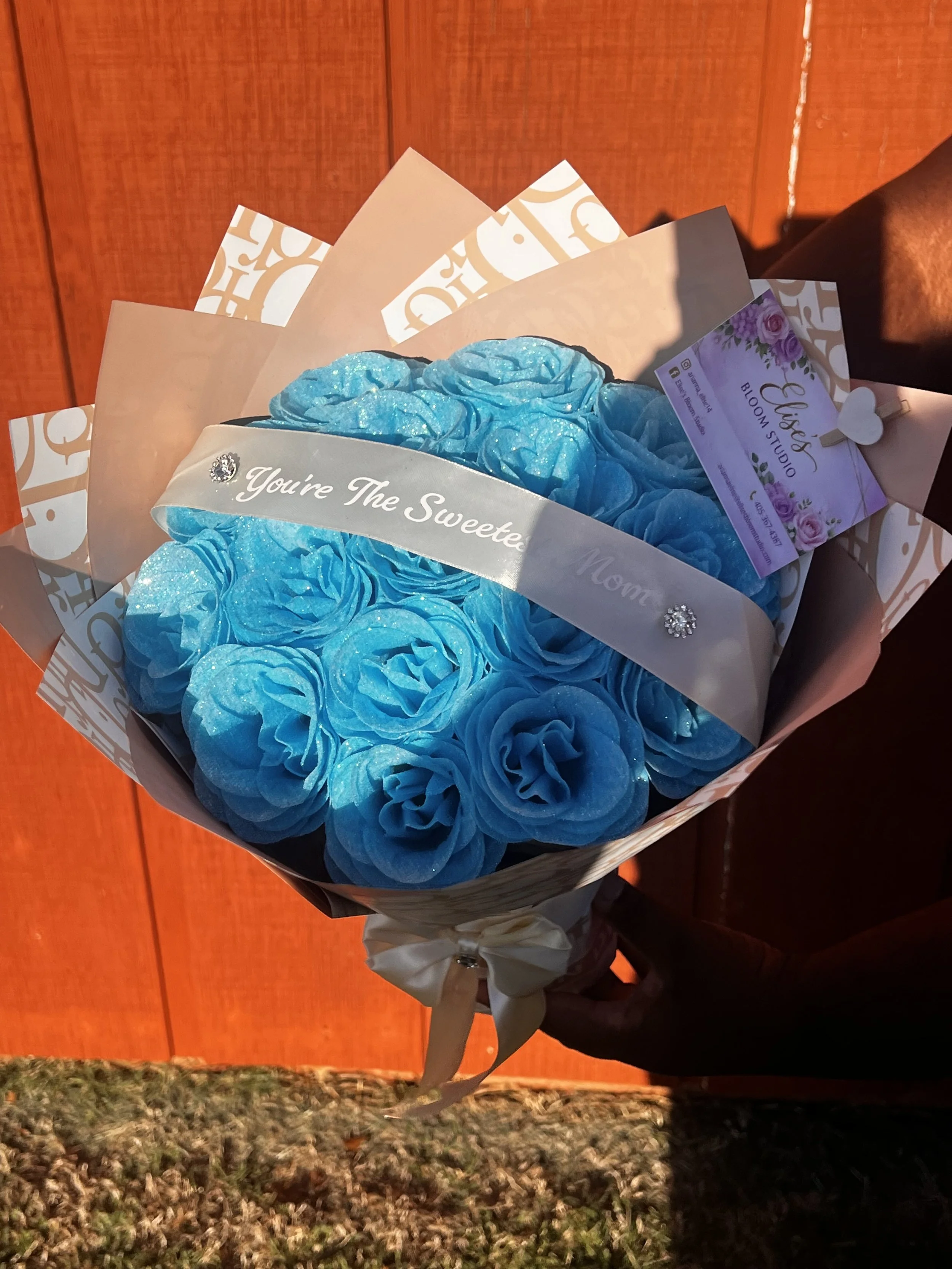 Hand holding a bouquet of blue roses wrapped in white and beige paper with a banner that reads 'You're The Sweetest' and a business card from Bloom Studio.