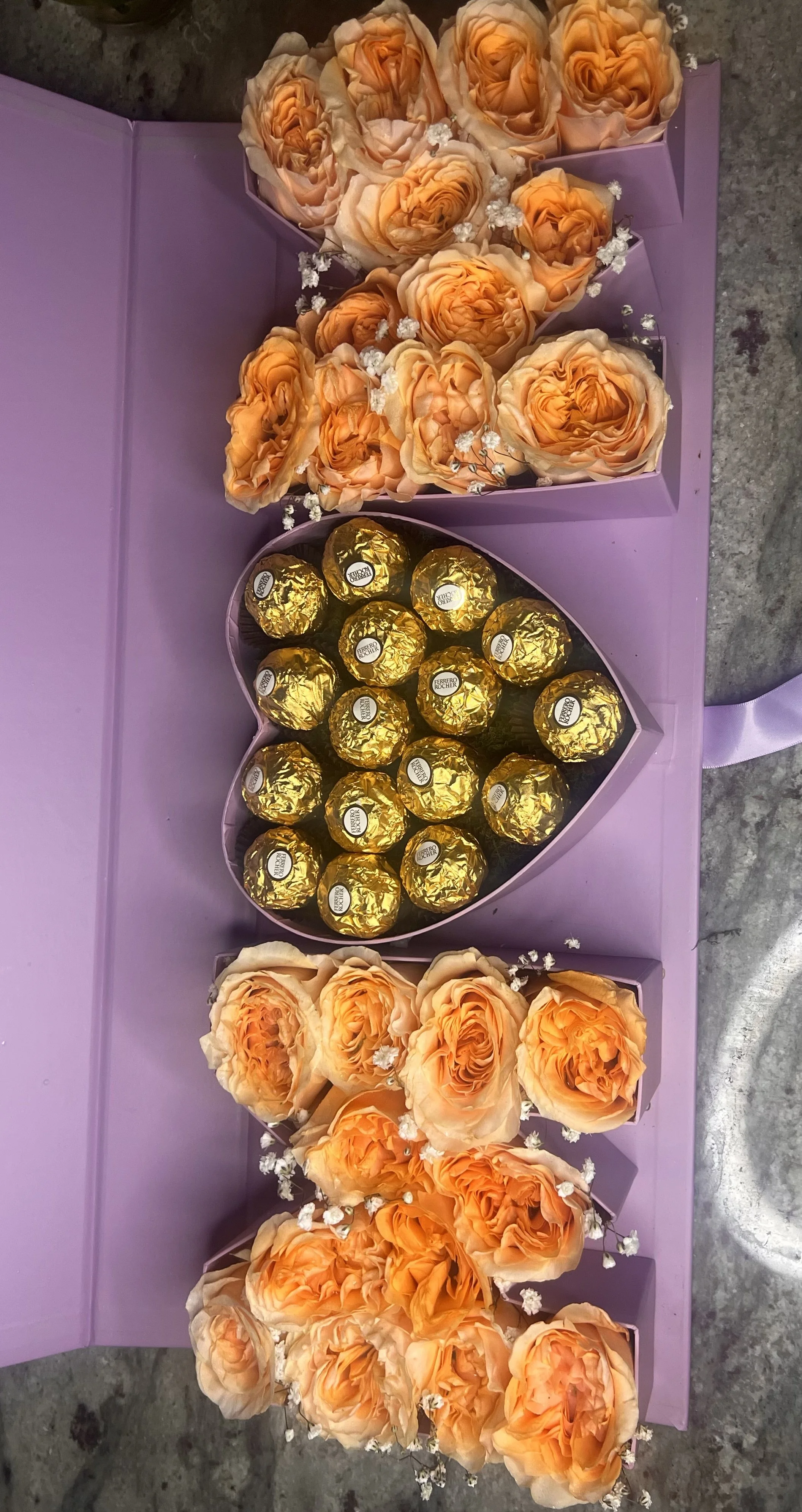 A purple gift box with peach-colored roses, and a heart-shaped box of Ferrero Rocher chocolates, decorated with small white flowers.