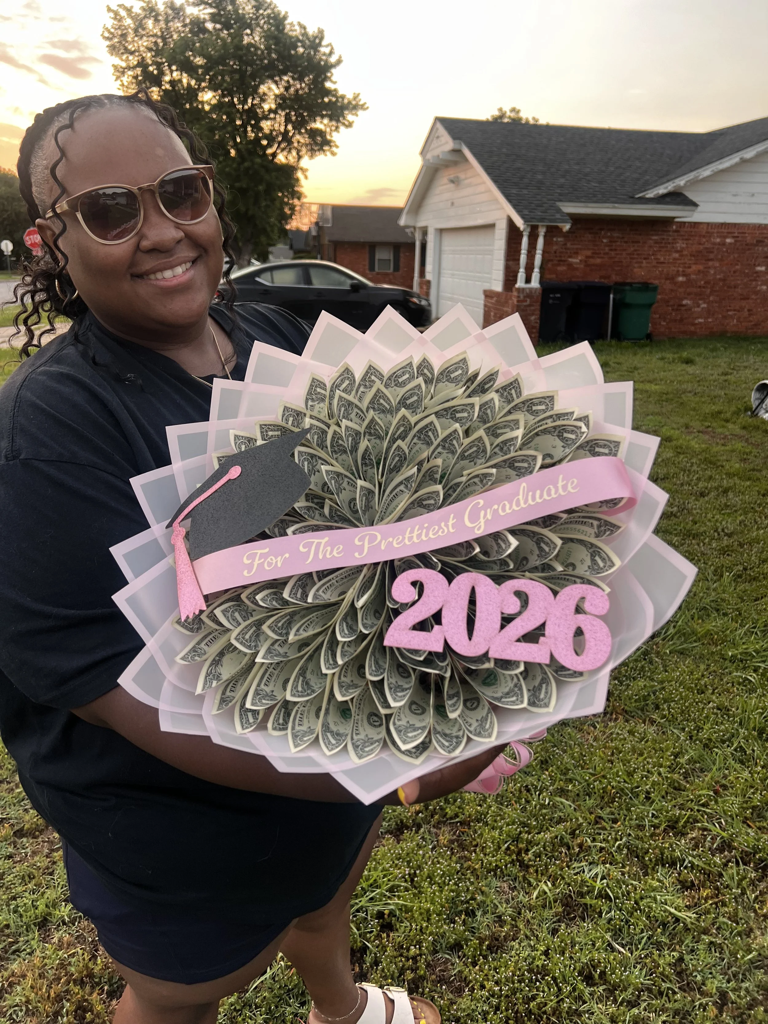 Woman holding a graduation bouquet made of folded dollar bills, decorated with a pink ribbon and sign that says 'For The Prettiest Graduate 2026,' wearing sunglasses, black shirt, and shorts, standing on a grassy area during sunset with a house and cars in the background.