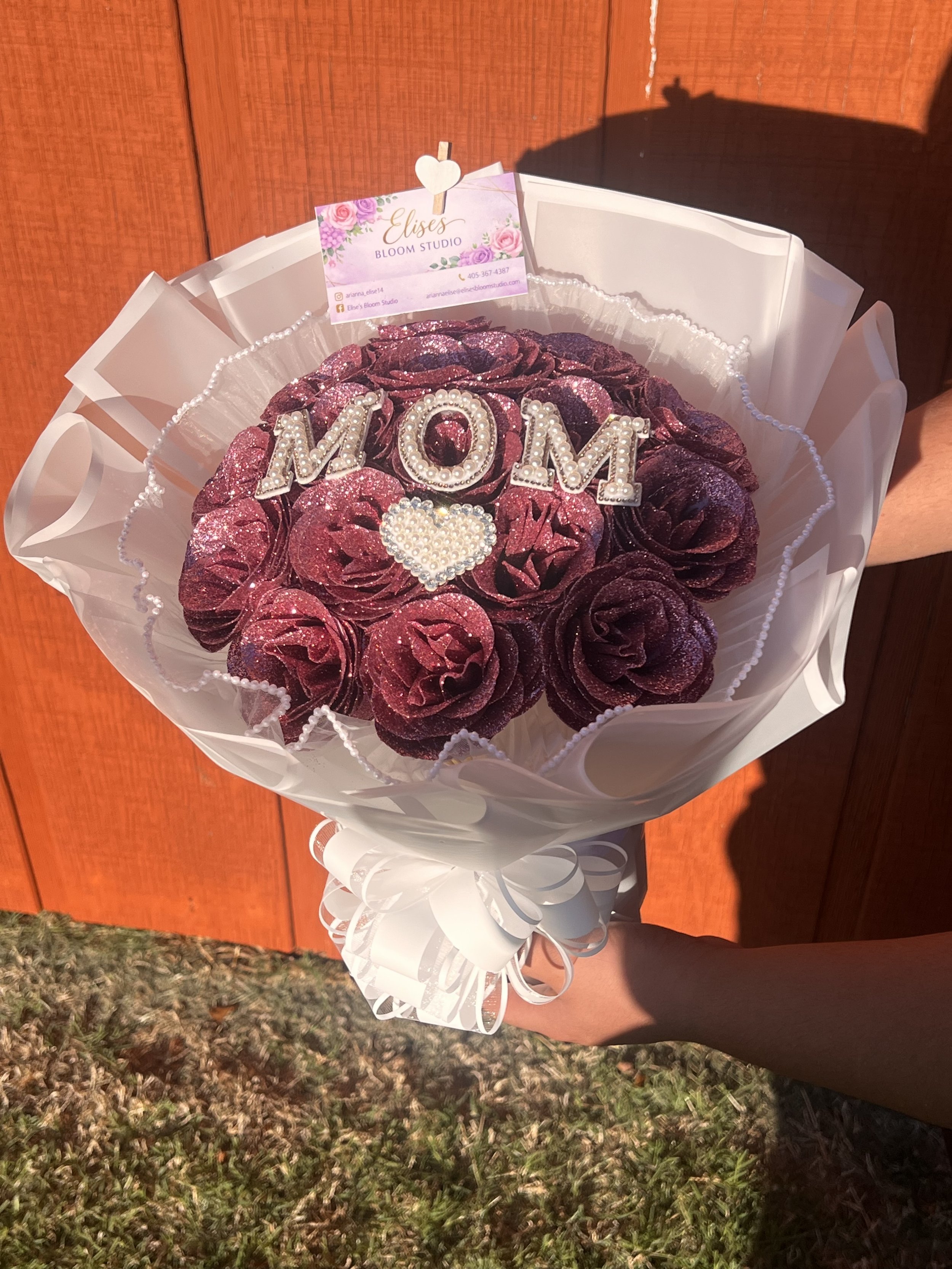 A bouquet of dark pink roses with glitter, decorated with the word 'MOM' in pearl and rhinestone letters and a pearl heart at the center, wrapped in white paper with a decorative string, and displayed against a wooden background.
