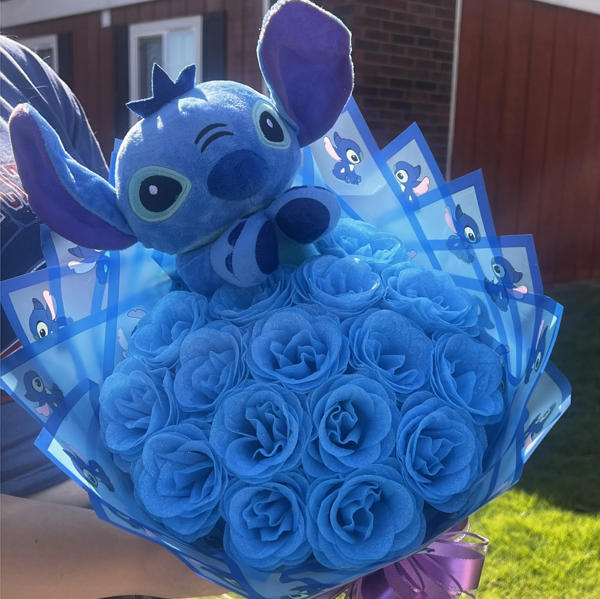 A large blue plush toy of the character Stitch from Disney's "Lilo & Stitch" is seen with a close-up of its face, surrounded by Blue-themed decorations including cards with images of Stitch and a bunch of blue paper or fabric, outside against a backdrop of a brick building and a grassy area.