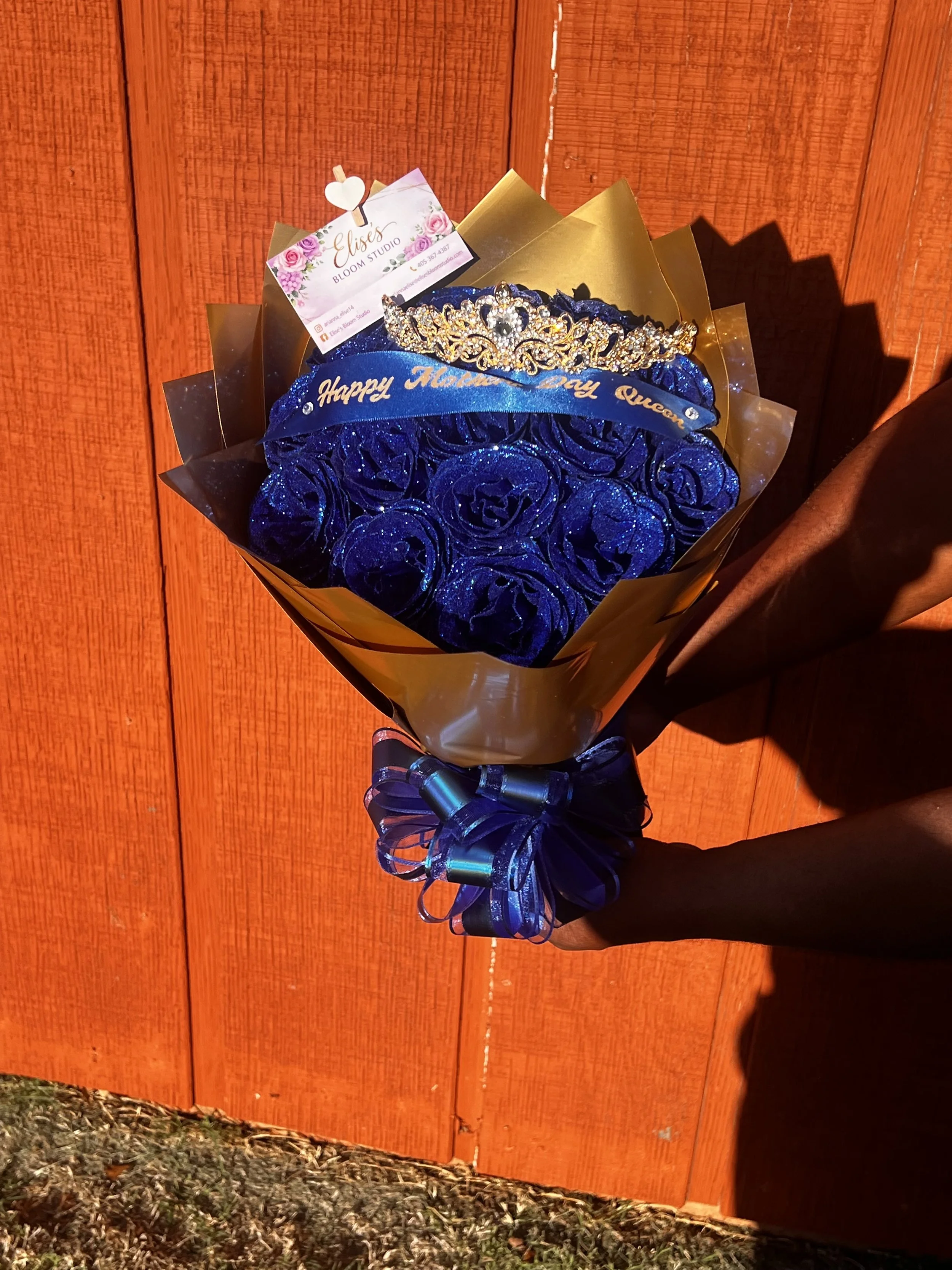 Bouquet featuring blue roses wrapped in gold paper, adorned with a blue ribbon that says "Happy Mother's Day Queen", topped with a gold tiara, and accompanied by a business card for Blossom Studio, held by a person with a dark sleeve against a wooden