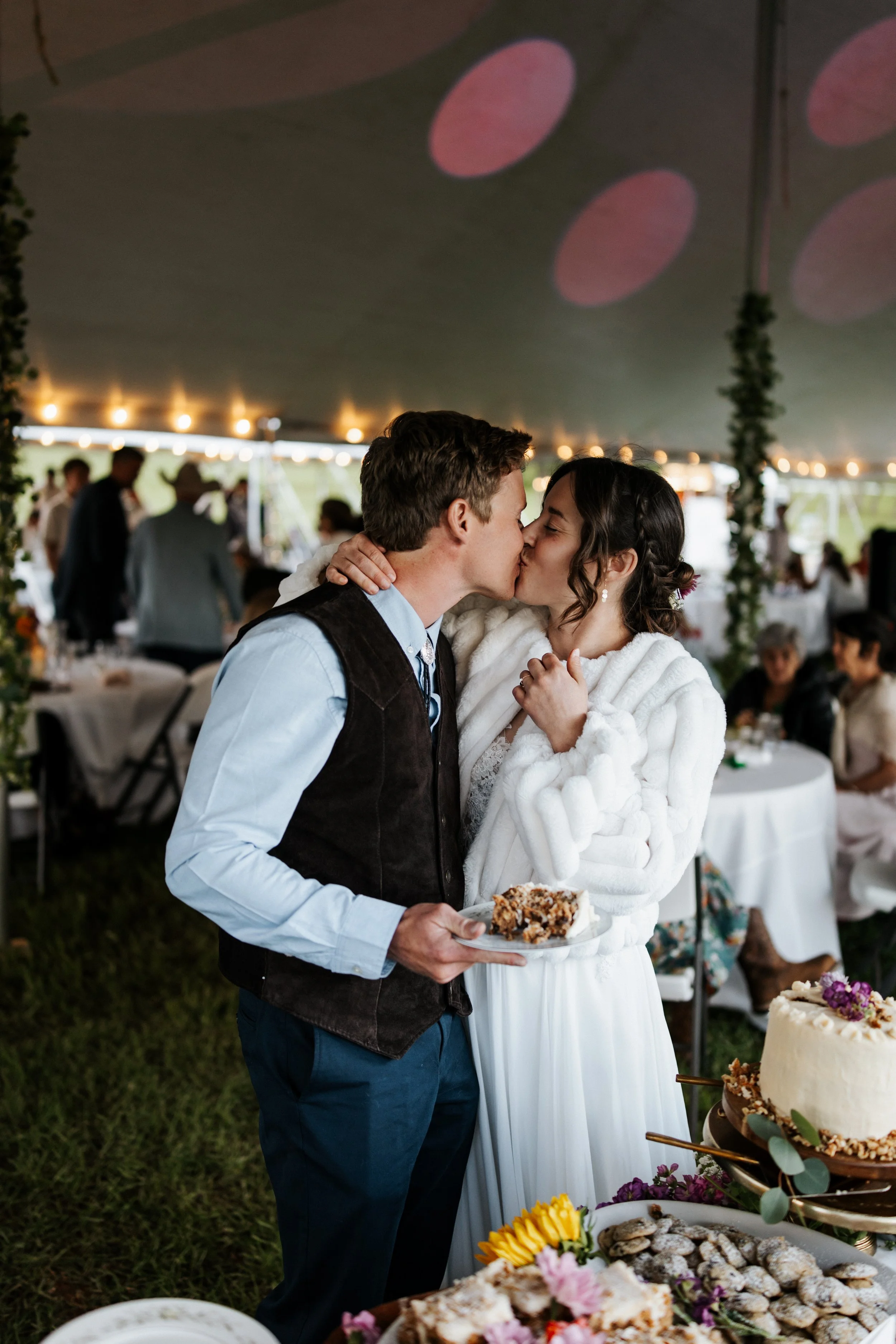 A couple sharing a kiss at a wedding reception under a tent, with tables of food and guests in the background.