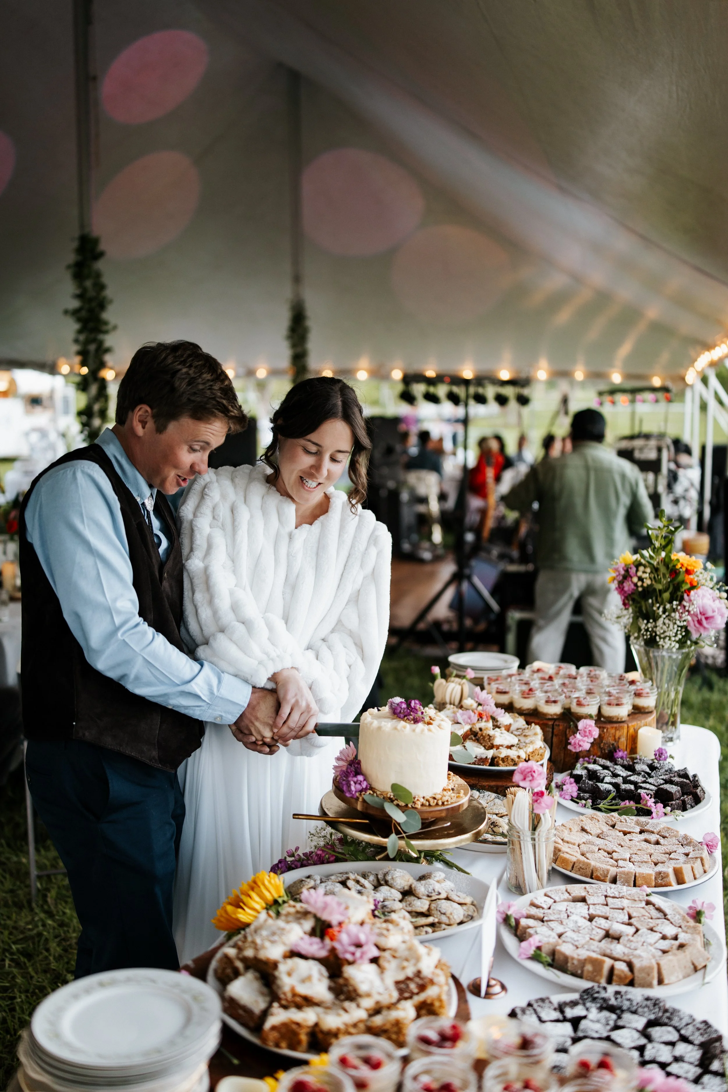 A couple is cutting into a wedding cake at an outdoor reception, surrounded by desserts and floral decorations under a large tent.
