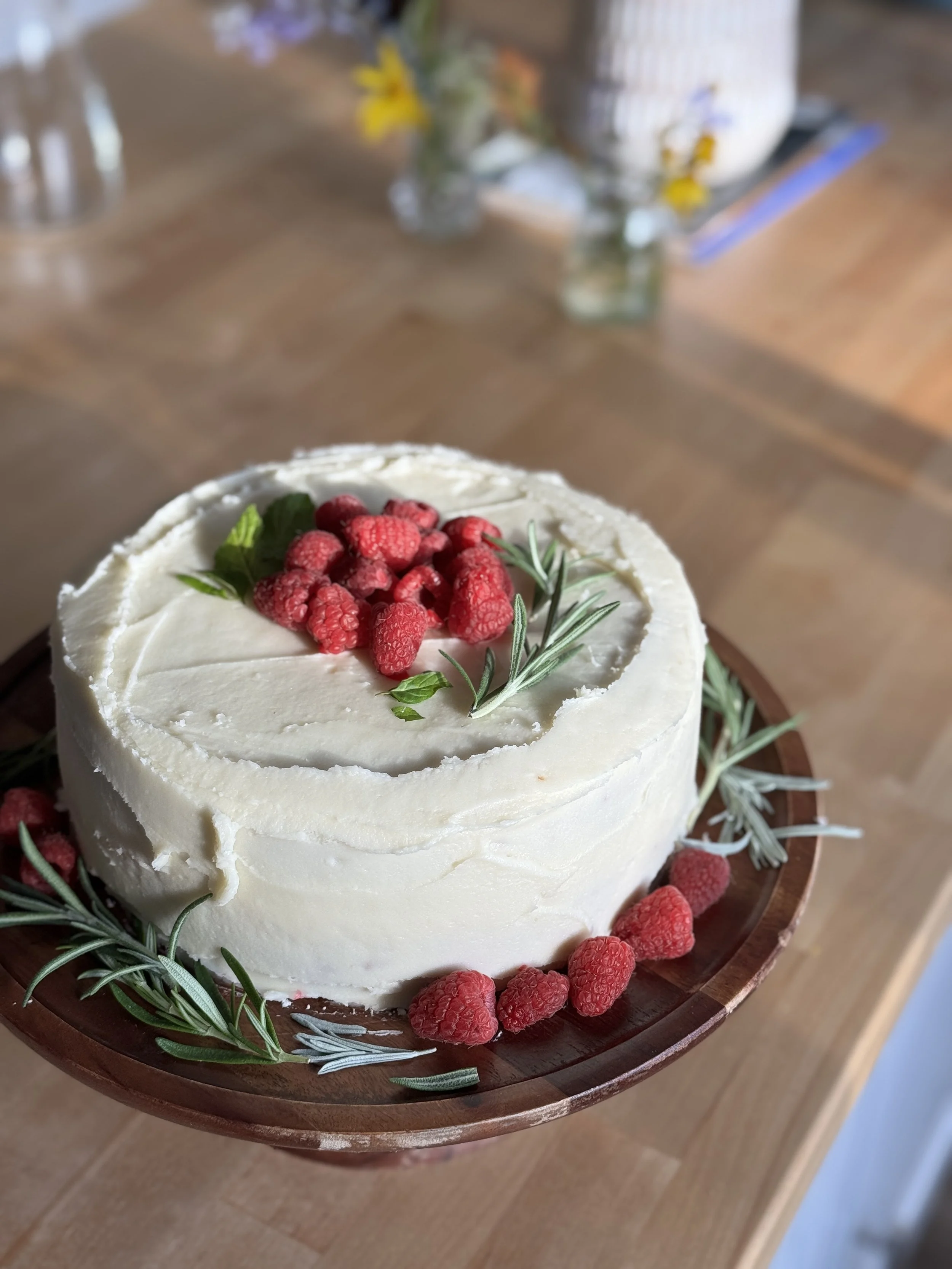 A round white frosted cake decorated with fresh raspberries and green herbs, surrounded by sprigs of rosemary, placed on a wooden serving tray.