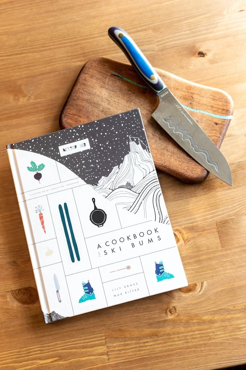 A cookbook titled 'A Cookbook for Ski Bum' by Lily Krass and Max Ritter placed on a wooden surface, next to a small wooden cutting board with a unique serrated knife that has a blue, purple, and white marbled handle.