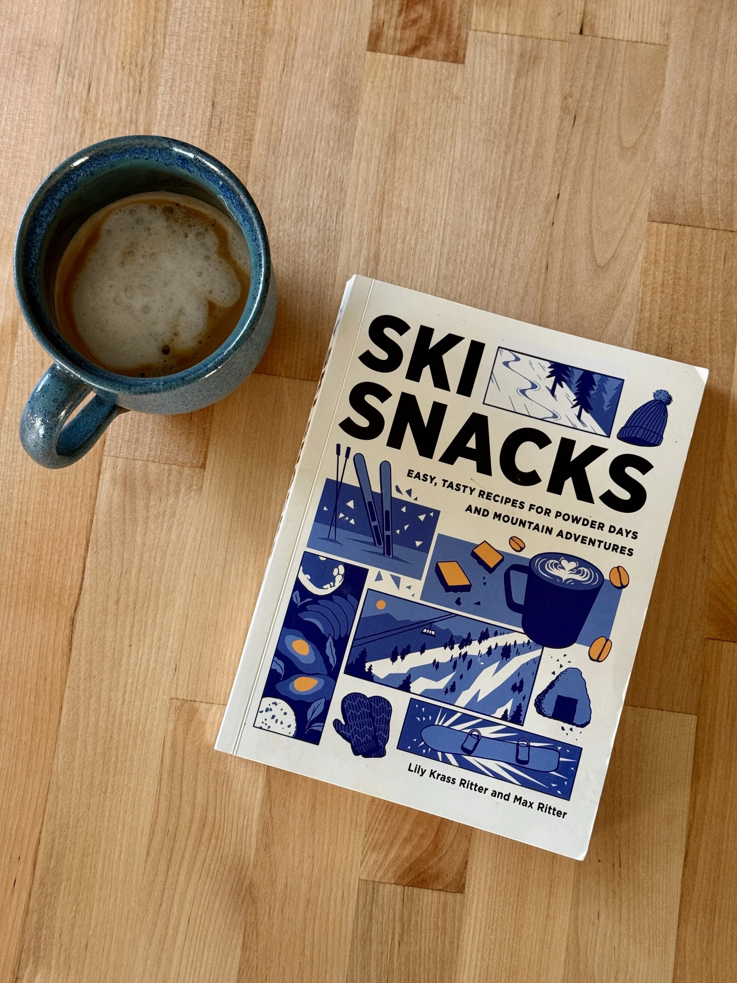 A blue ceramic mug filled with a frothy beverage sits on a wooden surface. Next to the mug is a book titled 'Ski Snacks' with a cover illustration of a winter landscape, ski equipment, and snacks.