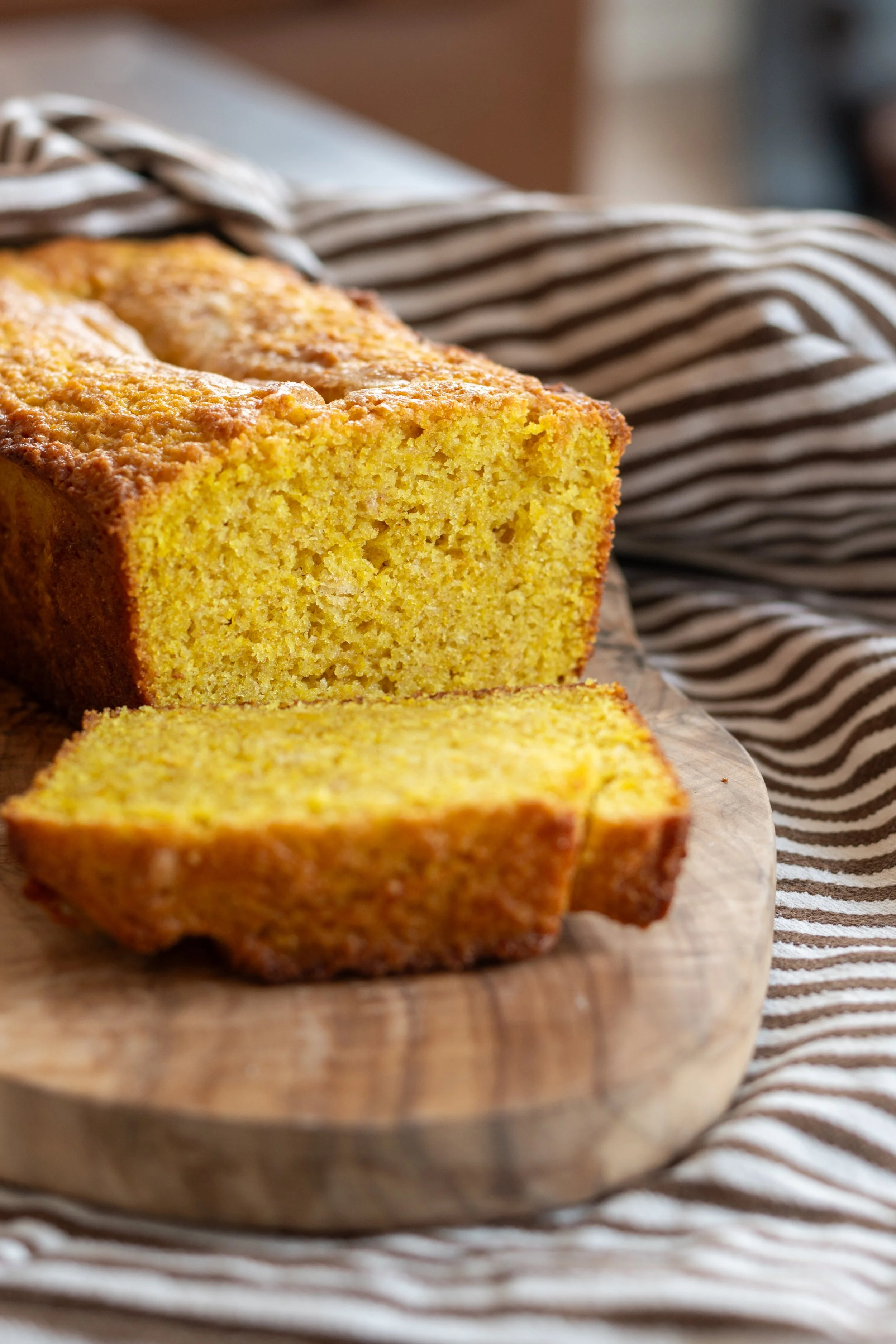 A freshly baked yellow yogurt cake loaf on a wooden cutting board, with a slice cut out, resting on a striped cloth.