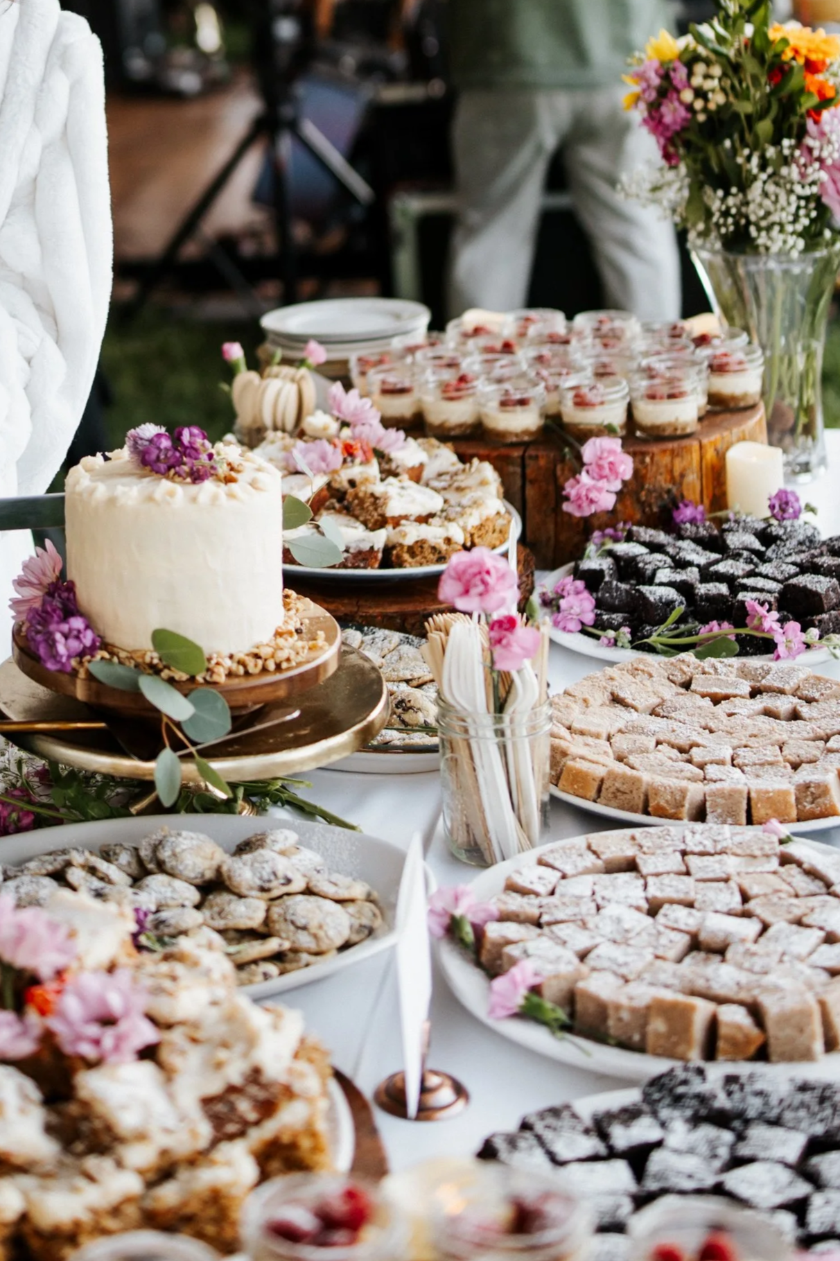 Variety of baked goods and desserts on a table, decorated with pink flowers, including a white frosted cake, cookies, brownies, and parfaits in small jars in the background.