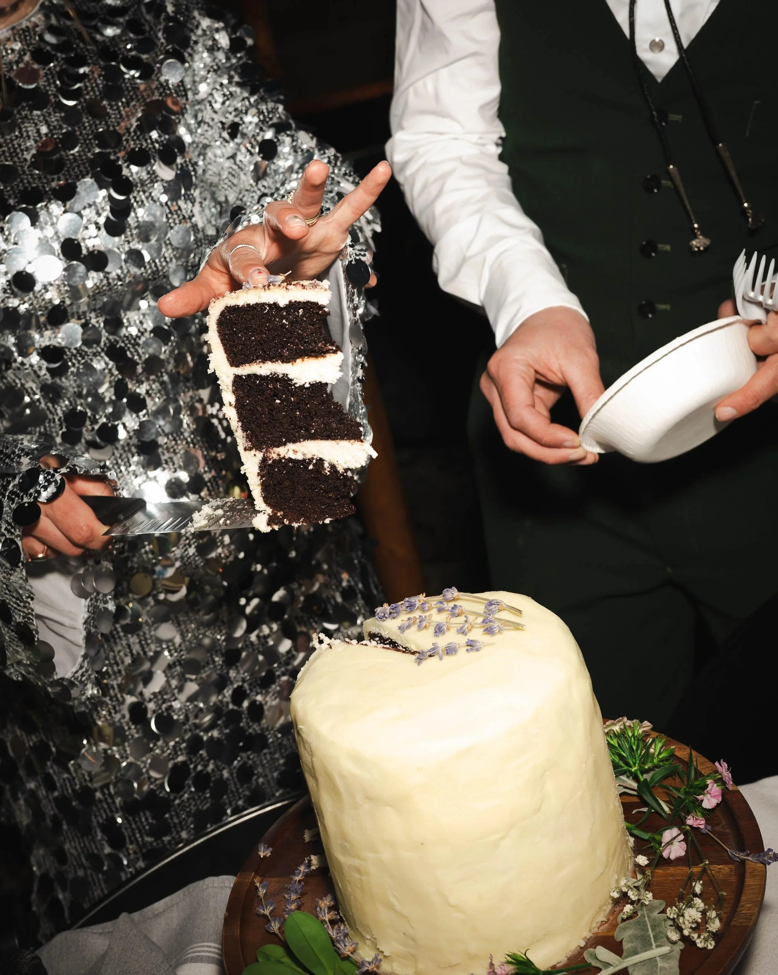 A person in a shiny, sequined outfit holding a large slice of chocolate cake with white frosting. The cake is decorated with small lavender flowers and surrounded by greenery and flowers on a wooden surface. Another person, partially visible, holds a white bowl and a fork.
