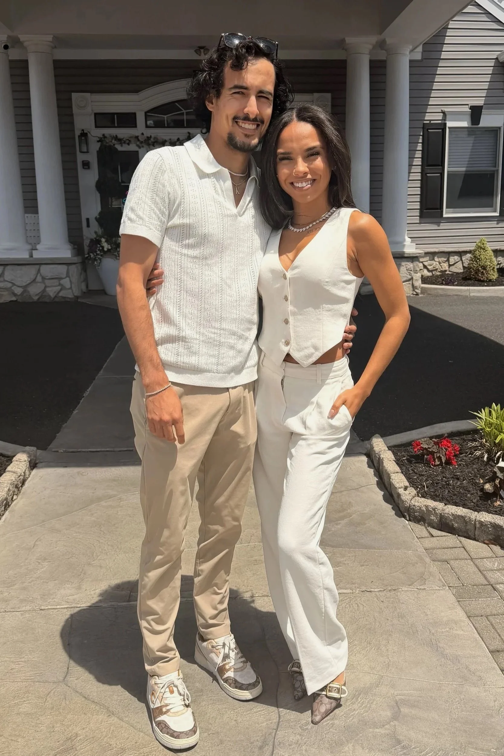 A man and woman standing outside in front of a house, smiling and posing for the photo. The man has dark, curly hair and a goatee, wearing a white short-sleeve shirt and beige pants. The woman has dark, shoulder-length hair, wearing a sleeveless white top with buttons and white pants. The house behind them is gray with white columns, windows, and flower beds.