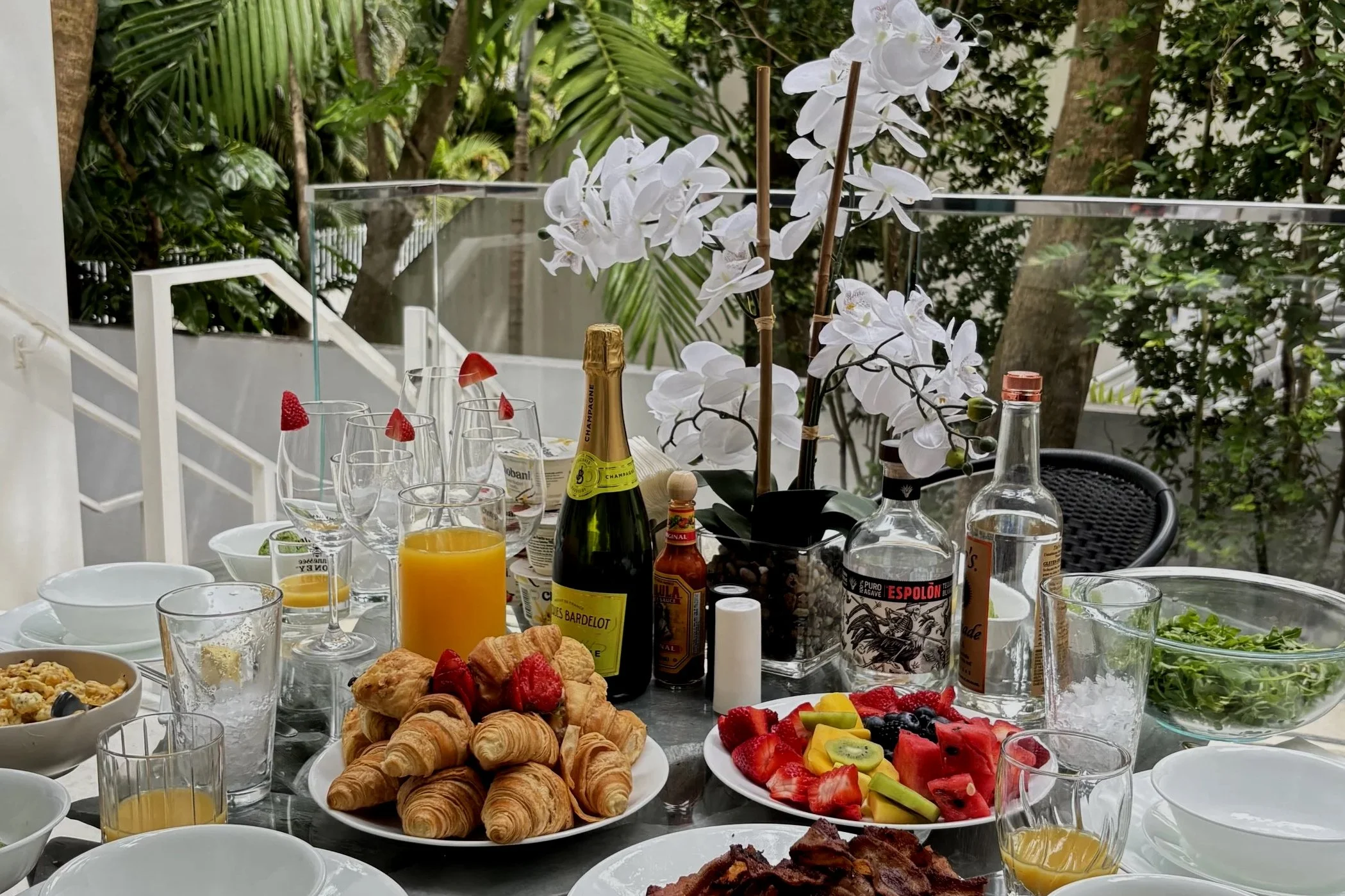 A table set for a meal with various foods and drinks, including croissants, pastries, fruit, juice, soda, and champagne, with white orchids and greenery in the background.