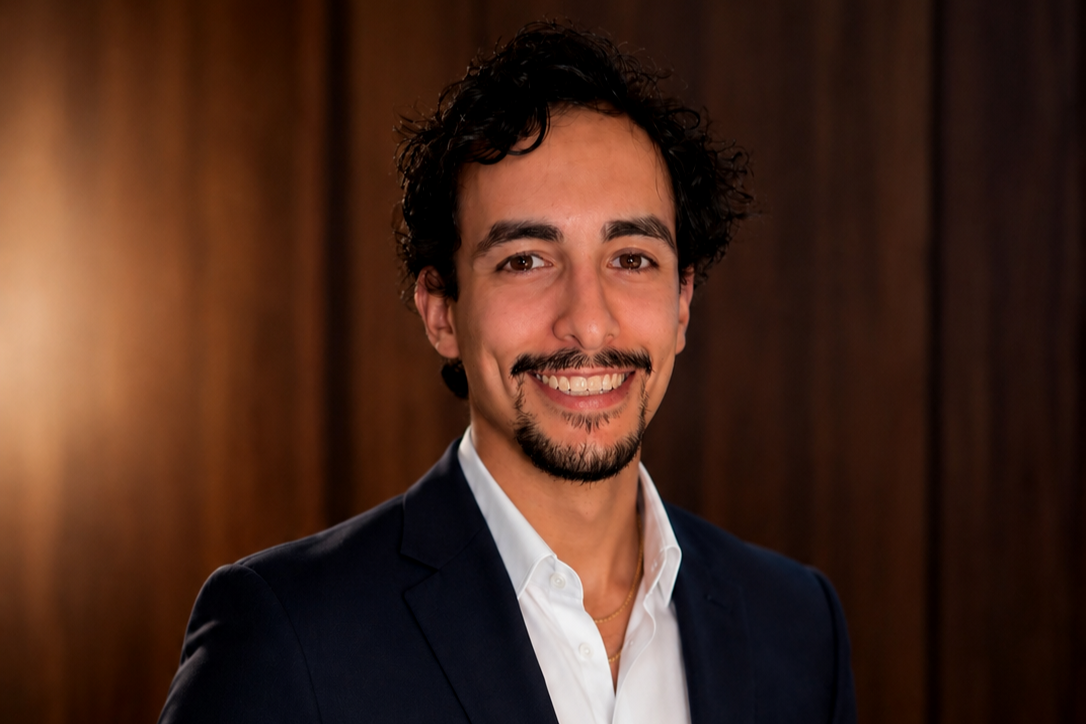 A man with dark curly hair, a goatee, and a mustache smiling in front of a wooden wall, wearing a white collared shirt and a dark blazer.
