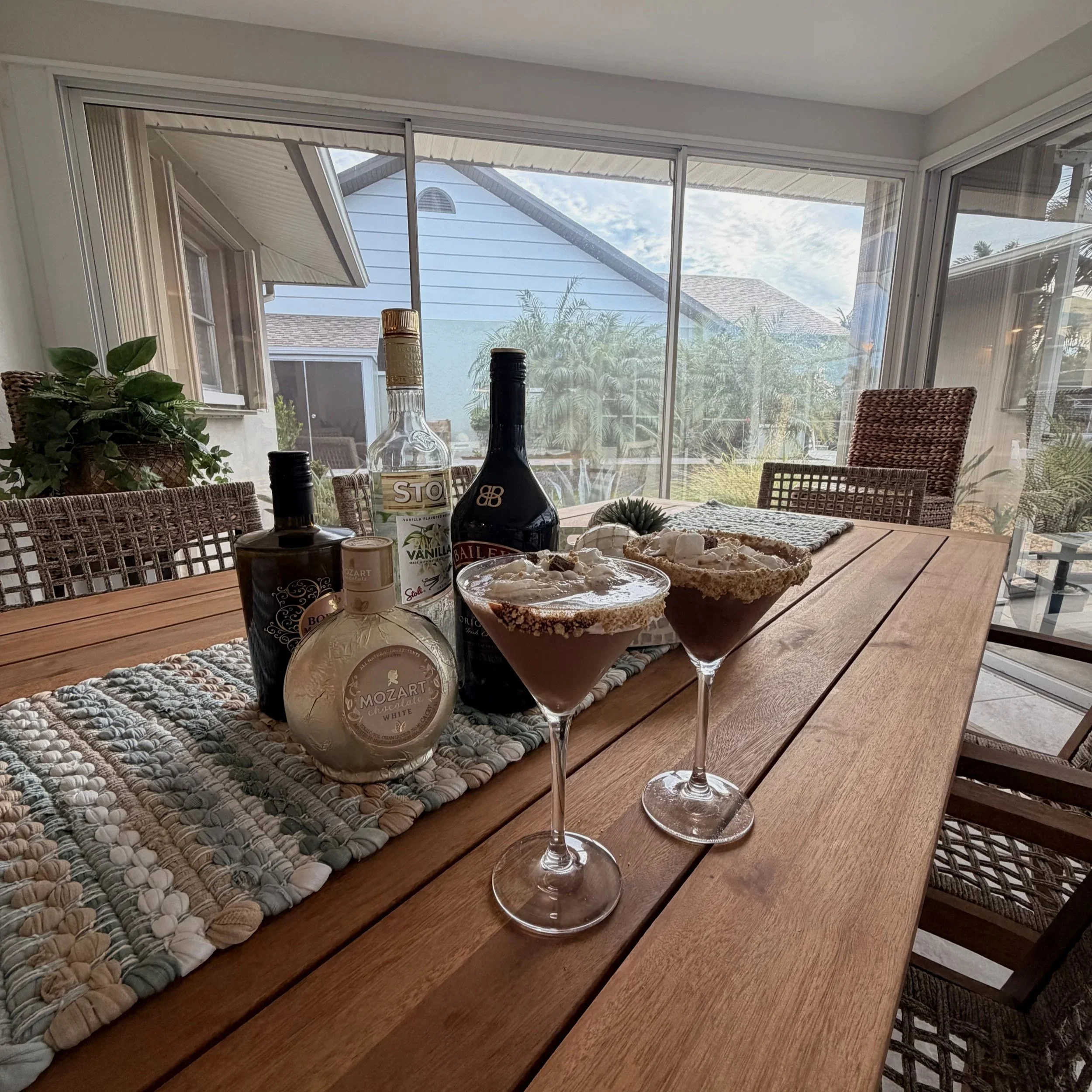 View of a dining area with a wooden table, wine bottles, cheese, and two empty wine glasses. The background shows a porch with outdoor furniture and a view of neighboring houses and trees through large glass sliding doors.