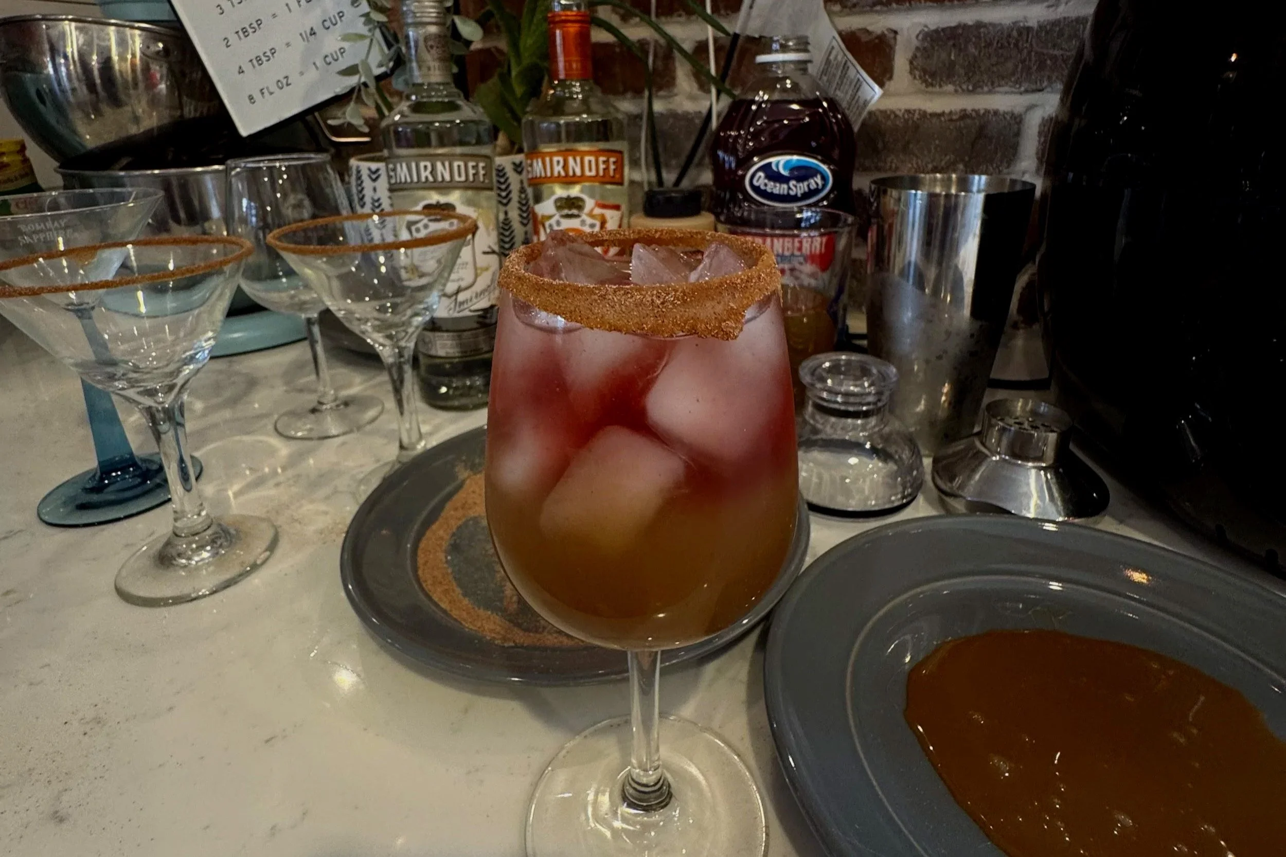 A cocktail glass with a salted rim, filled with ice and a reddish liquid, placed on a tray on a bar counter with various bottles, glasses, and bar tools in the background.
