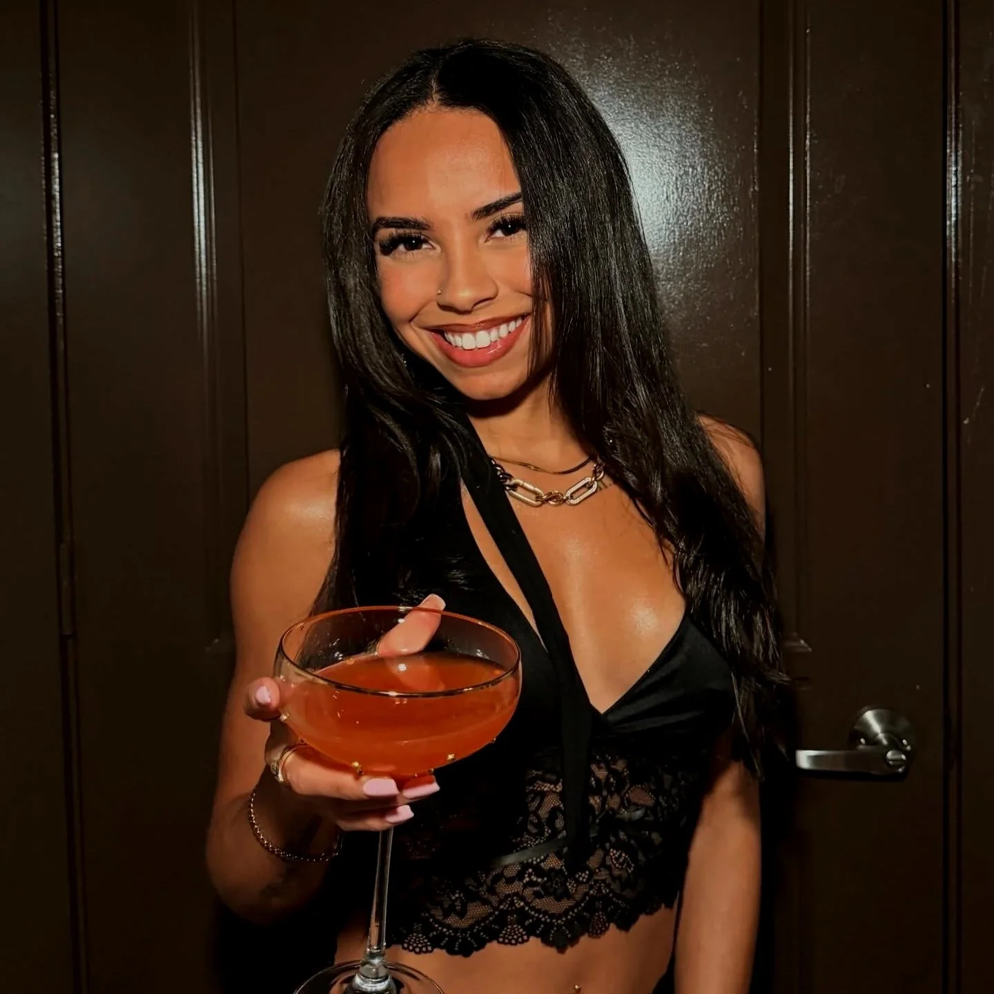 A woman with long dark hair holding a cocktail glass with an orange-colored drink, smiling, wearing a black lace top and jewelry, standing in front of a brown door.