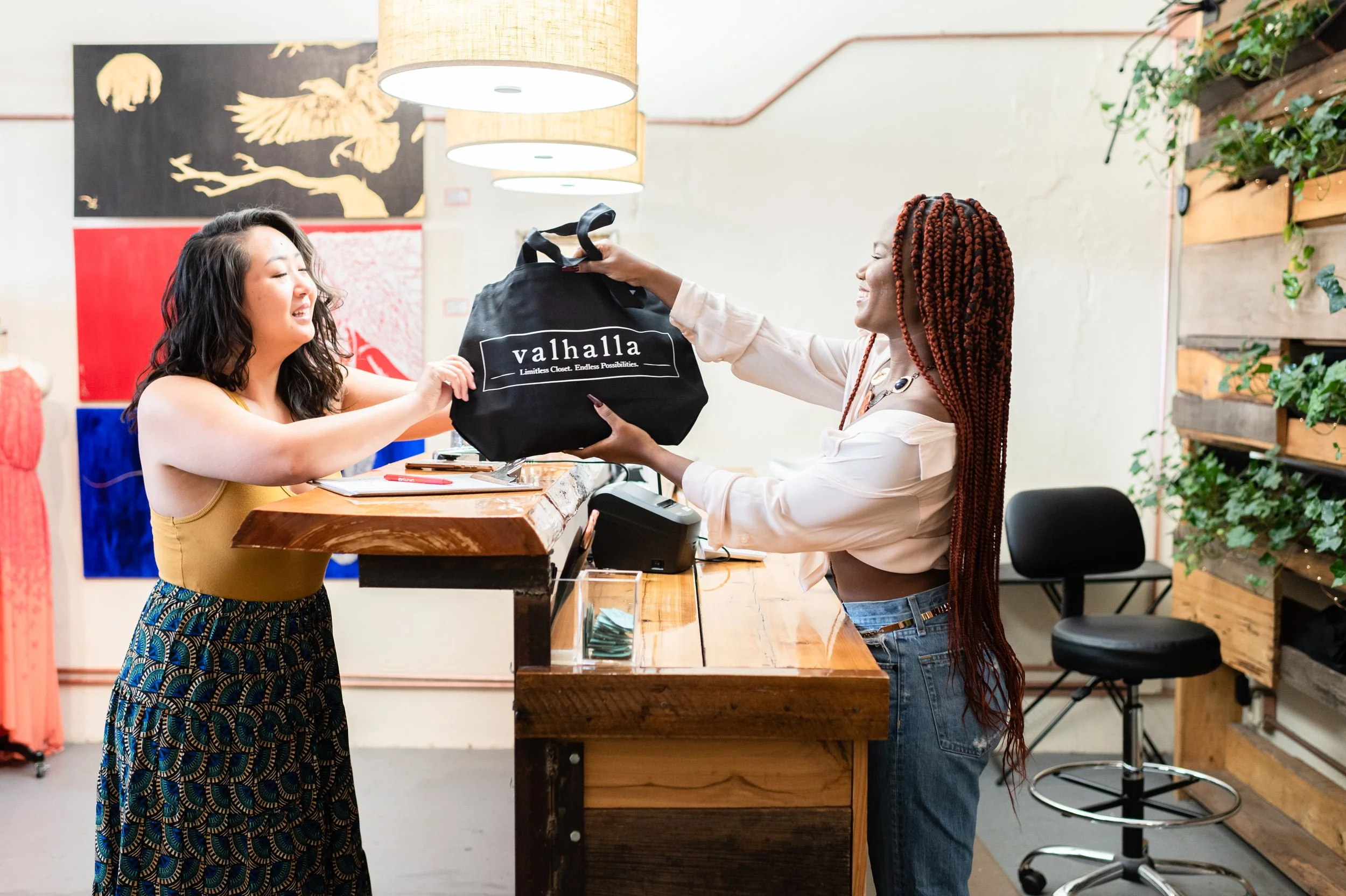 Two women at a retail store counter exchanging a black shopping bag with the logo 'valhalla' and slogan 'Limitless Closet. Endless Possibilities.' The woman on the left wears a yellow tank top and patterned pants, smiling, while the woman on the right, in a white blouse and jeans, smiles as she hands over the bag. The background features wall art, plants, and store decor.