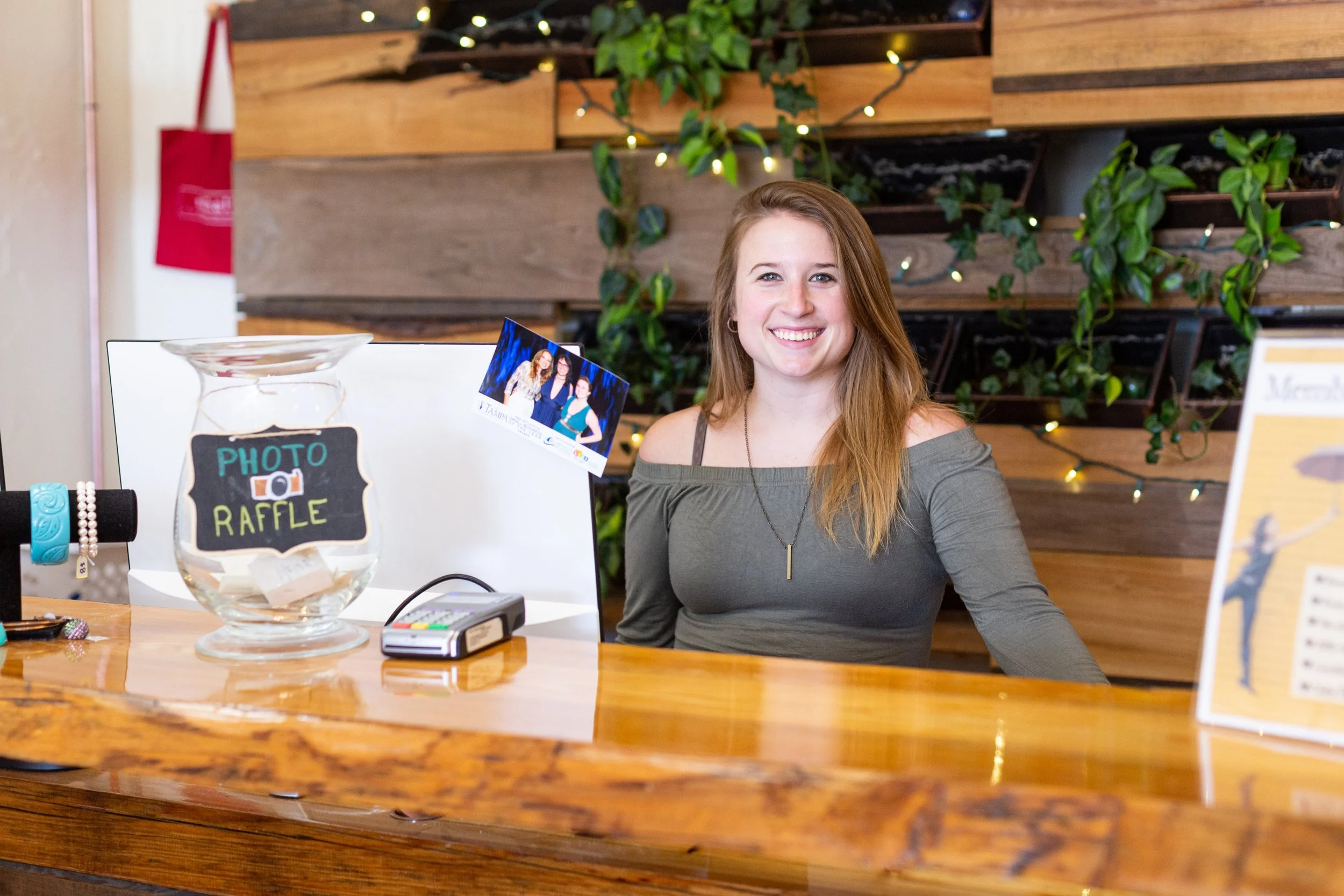 A smiling young woman with long red hair sitting at a wooden counter, with a photograph of herself and friends on the counter, and a glass jar labeled 'Photo Raffle' in front of her, in a warmly lit indoor space decorated with plants and string lights.