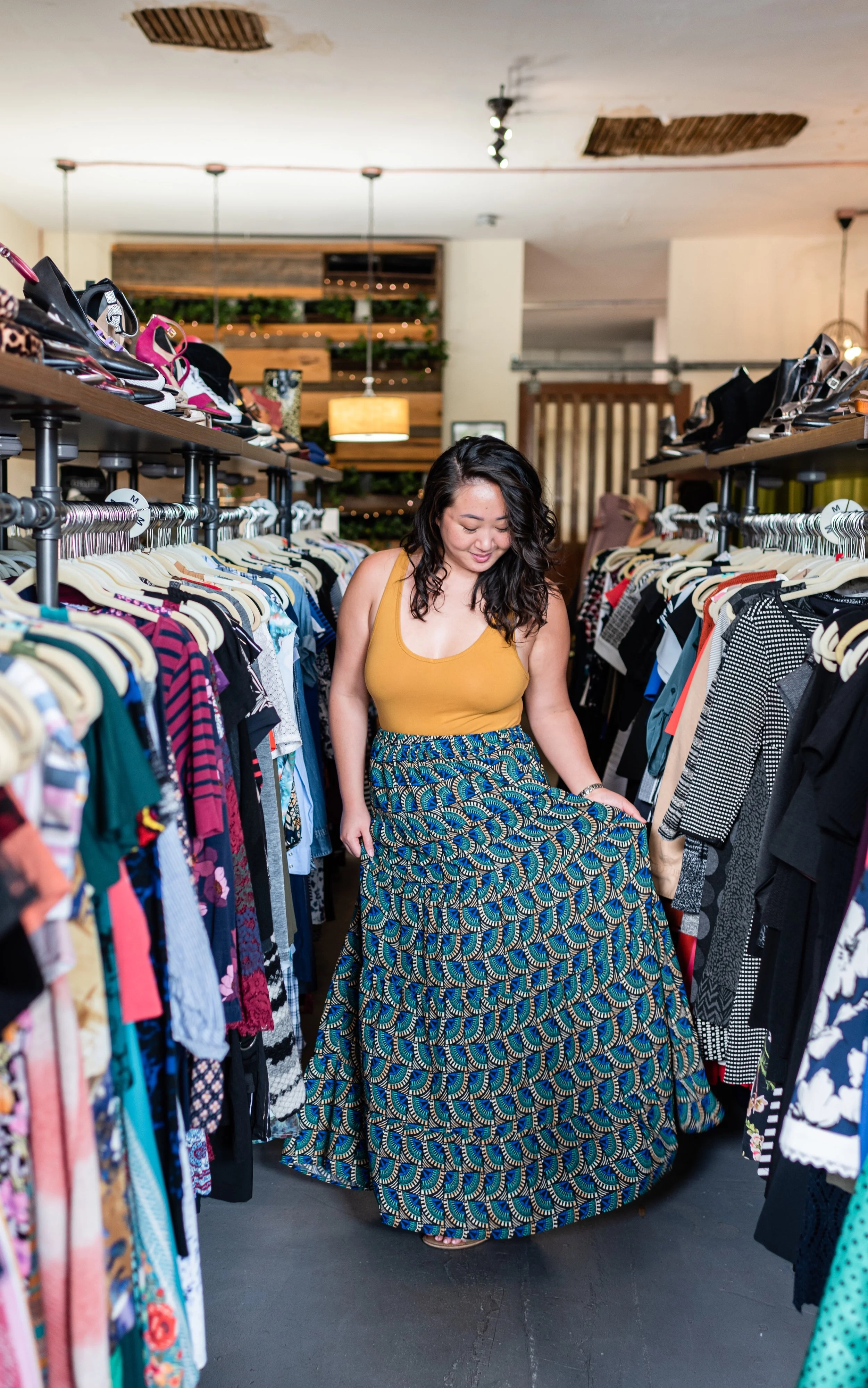 A woman shopping in a clothing store, holding a long colorful skirt while browsing racks of dresses and tops.