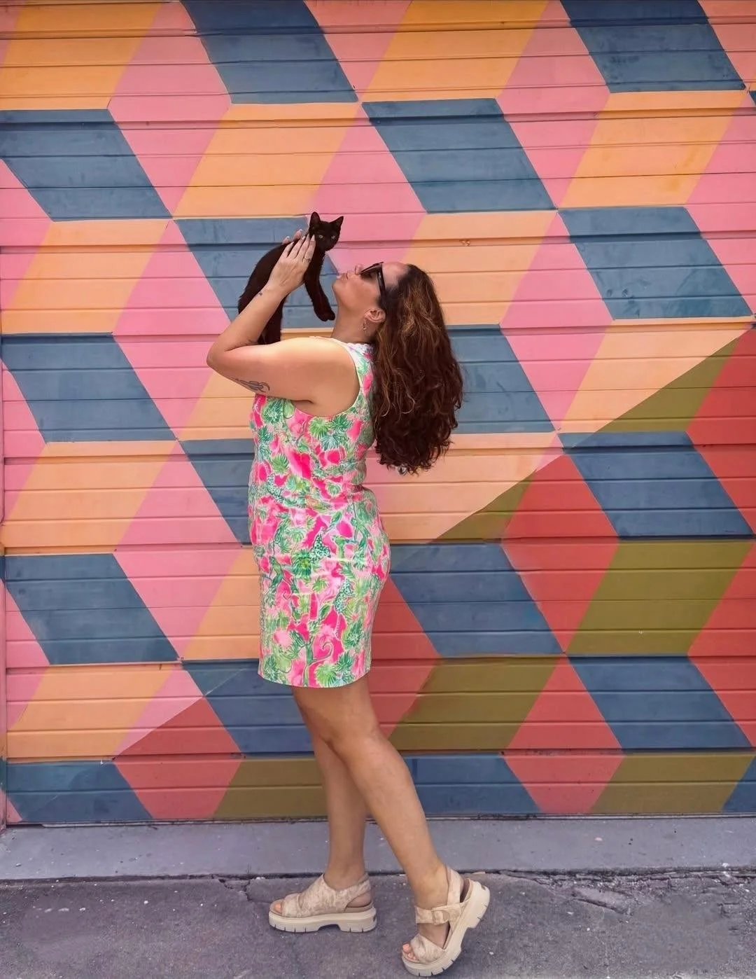 A woman with curly brown hair, wearing a colorful pink, green, and white dress, holds a small black cat up to her face against a vibrant, geometric painted wall.