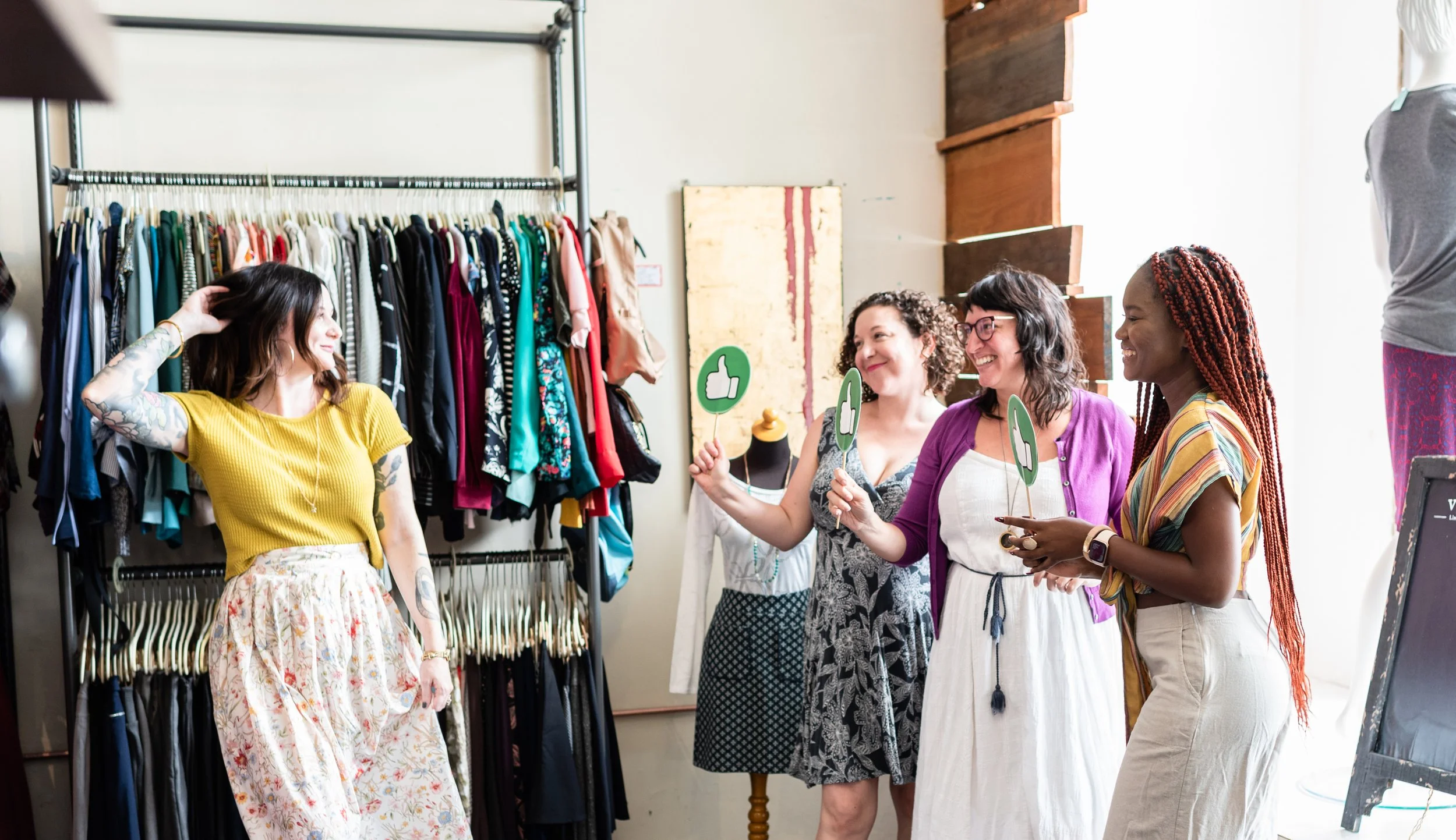 A woman with tattoos and wavy brown hair in a yellow top and floral skirt is talking to a group of women in a clothing store. The women are holding green thumbs-up signs. The store has a clothing rack filled with various dresses and colorful garments.