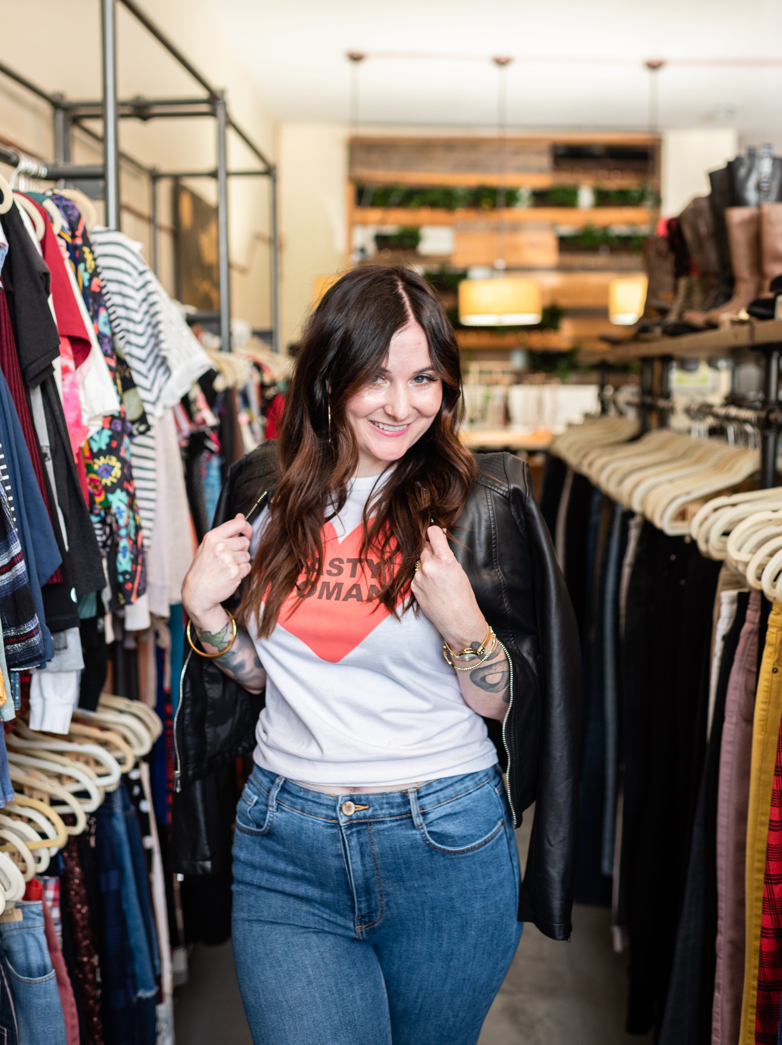 A woman with long brown hair and tattoos on her arm, smiling and wearing a black leather jacket over a white t-shirt with a red heart logo, standing in a clothing store aisle surrounded by racks of clothes.