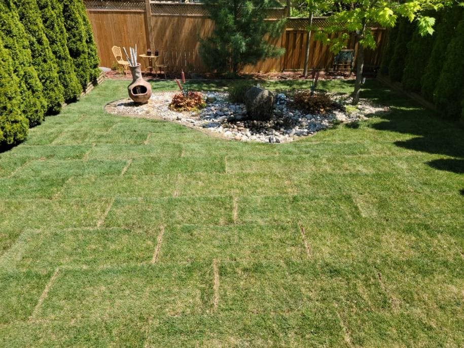 A backyard with well-maintained grass and a small landscaped area with rocks, plants, and trees. There is patio furniture and a clay chiminea on the left side, with a wooden fence in the background.