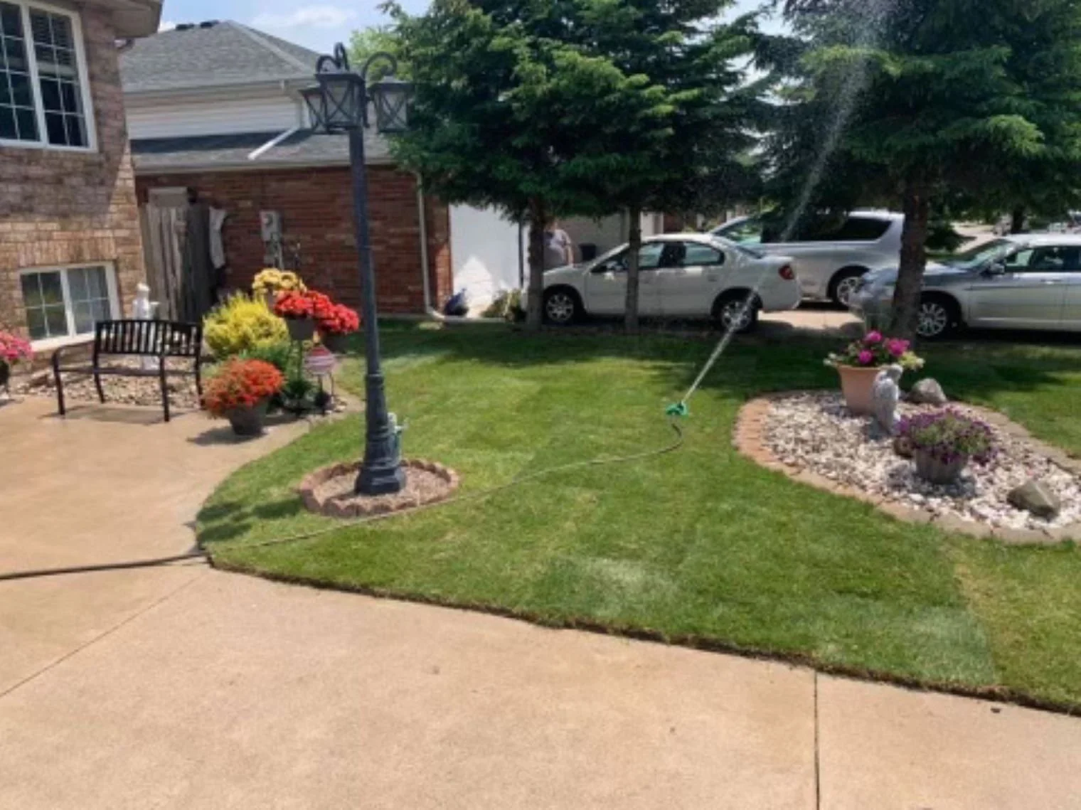 A well-kept backyard garden with a green lawn, flower beds with potted flowers, a bench, a street lamp, and cars parked in the driveway. A sprinkler is watering the grass, and trees provide shade.